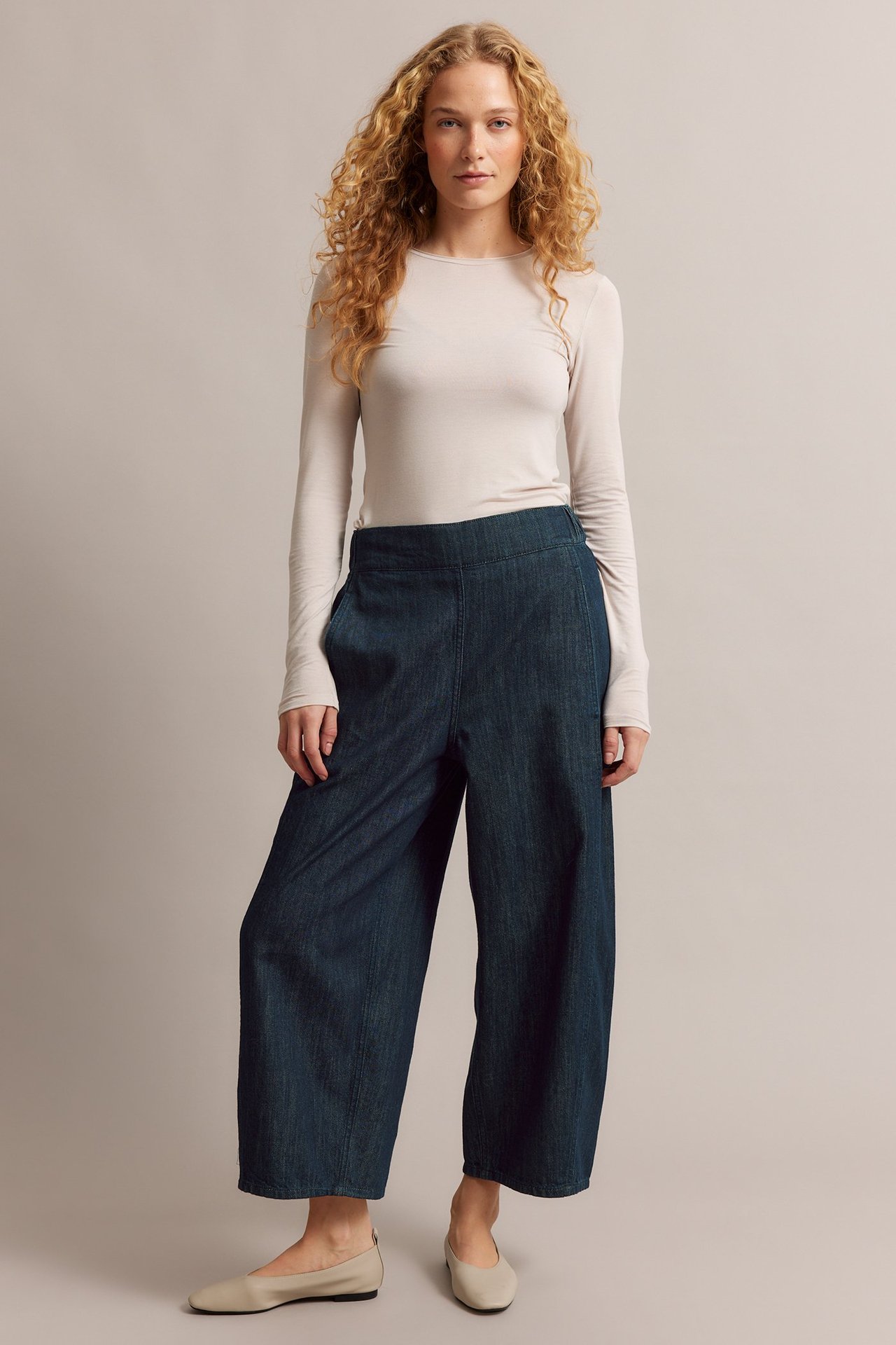 Edestä: Nainen tummansinisissä leveissä farkuissa, vaaleabeigessä pitkähihaisessa paidassa ja beigeissä pikkukengissä.