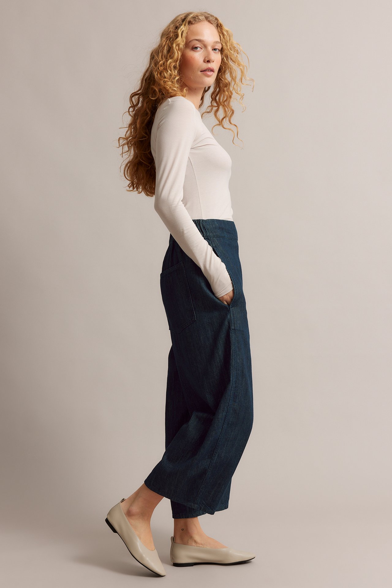 Widok z boku kobiety w ciemnych barrel leg dżinsach pull-on, jasnobeżowym topie z długim rękawem i jasnobeżowych balerinkach.