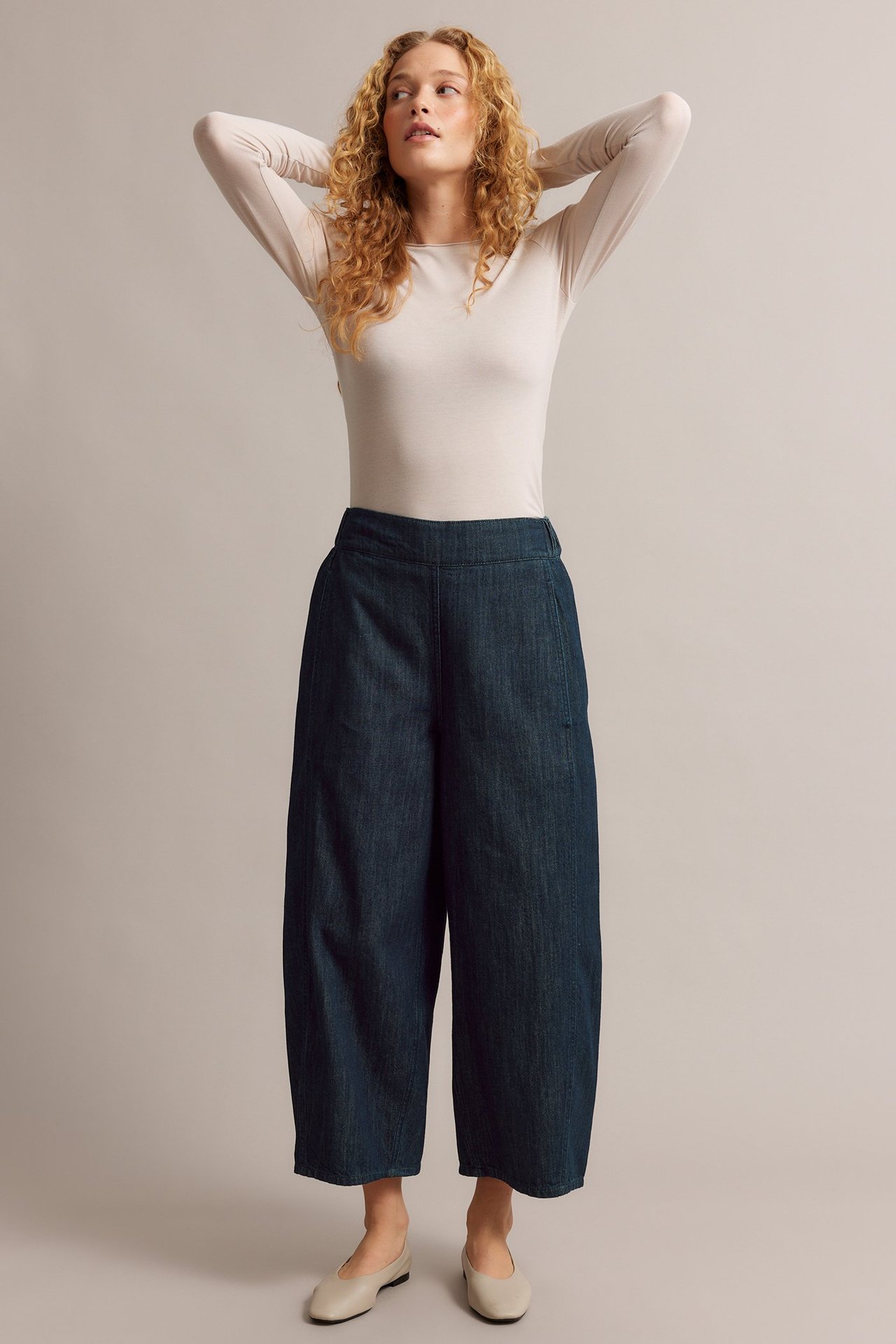 Edestä: nainen tummansinisissä barrel leg -pull-on-farkuissa, vaalea pitkähihainen toppi ja beiget avokkaat.