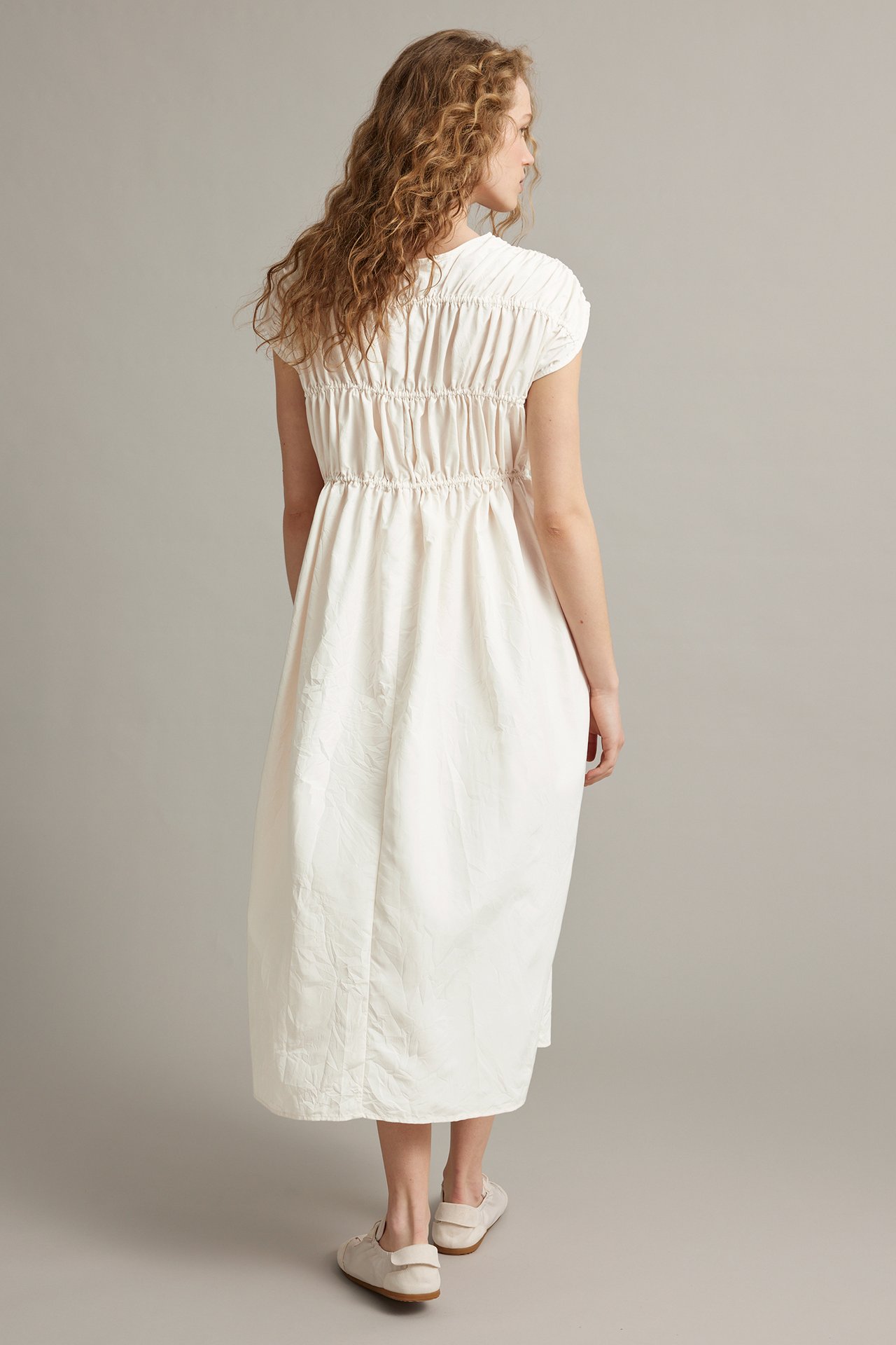 Naisen selkä, yllään valkoinen rypytetty midimekko lyhyillä hihoilla ja beiget matalat kengät.