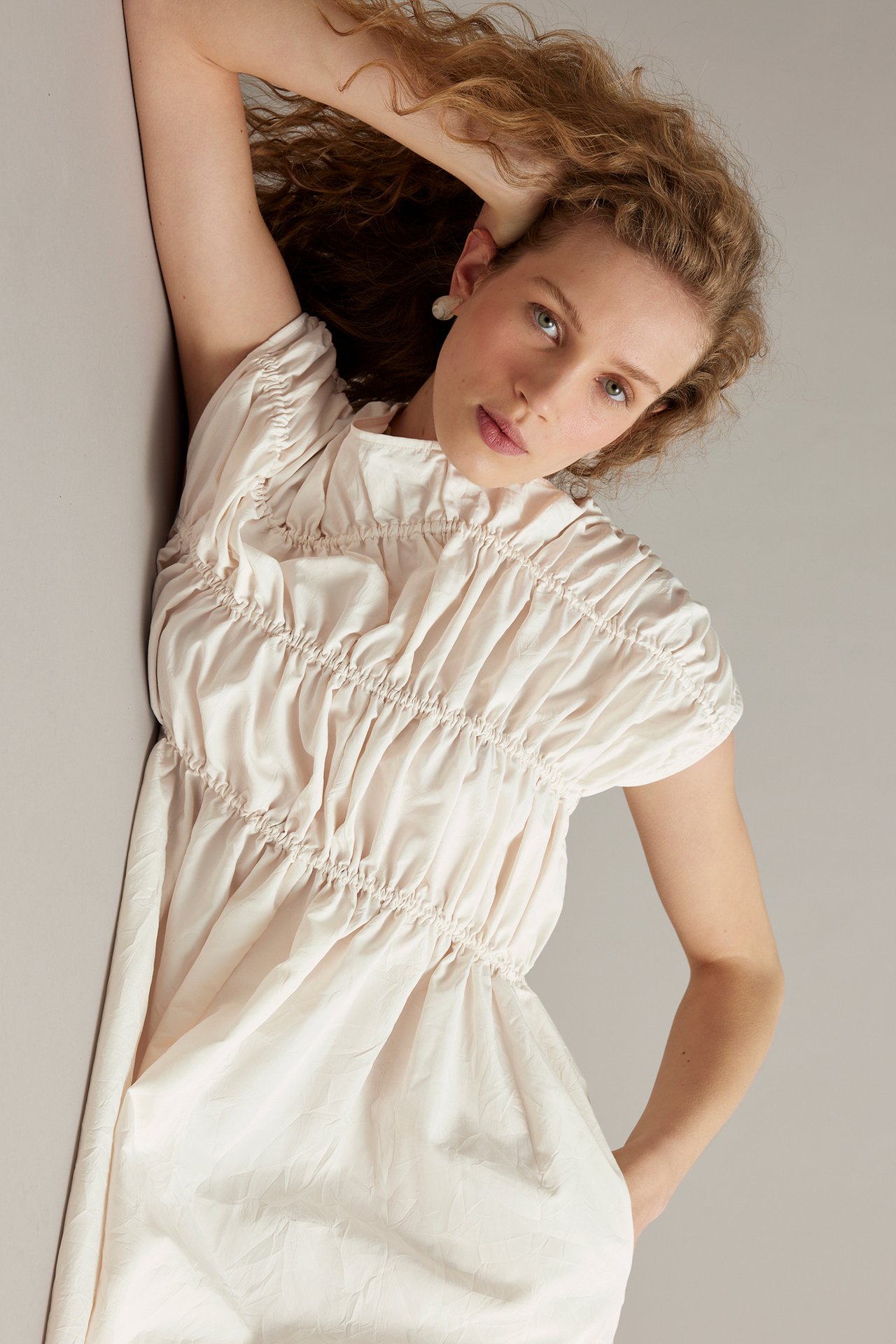 Zbliżenie: kobieta w białej, strukturalnej sukience z marszczoną górą i krótkimi rękawami, z perłowymi kolczykami.