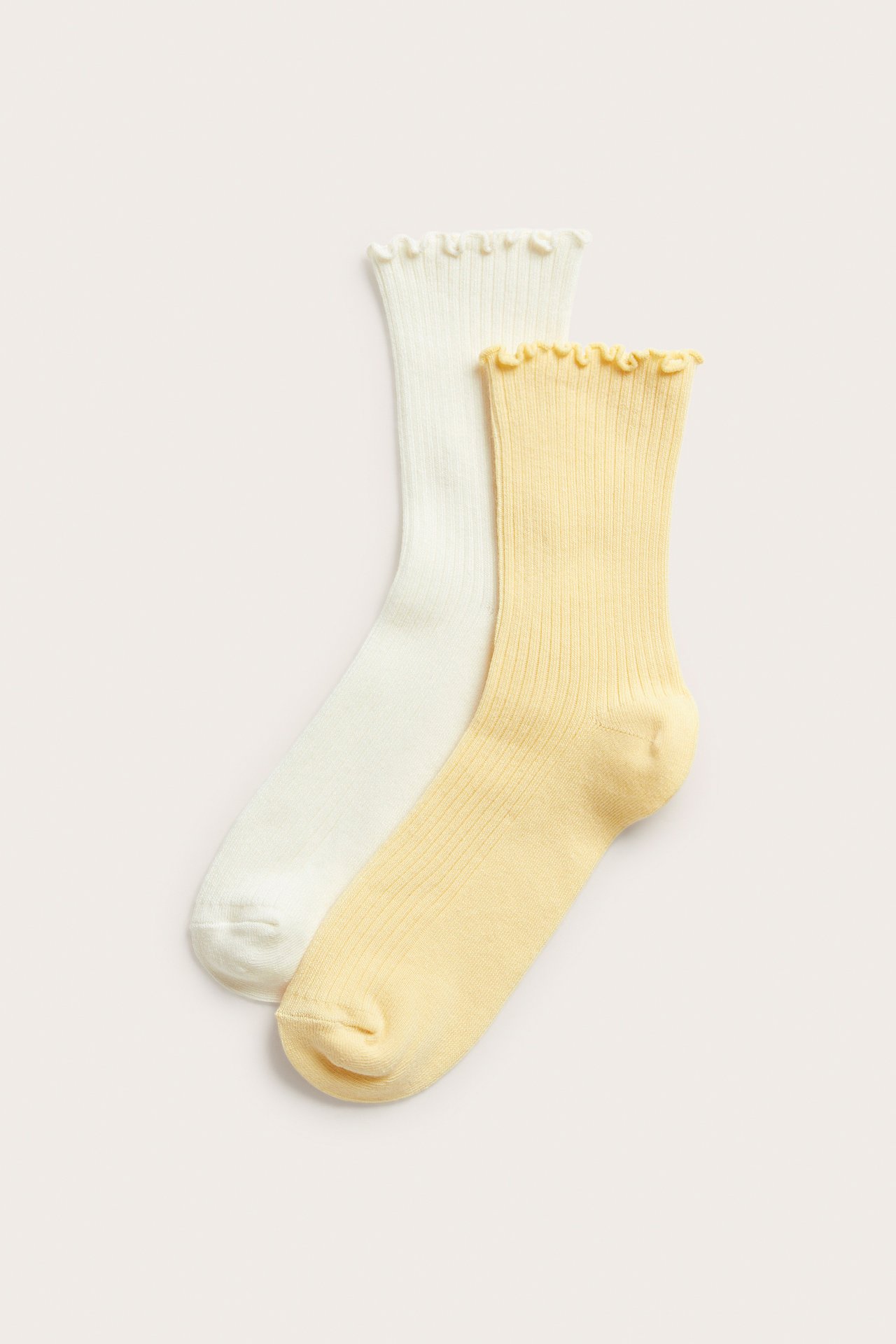 Edestä: Kaksi ribbineuletta sukkaa, toinen vaaleankeltainen, toinen luonnonvalkoinen, molemmissa röyhelöreuna.