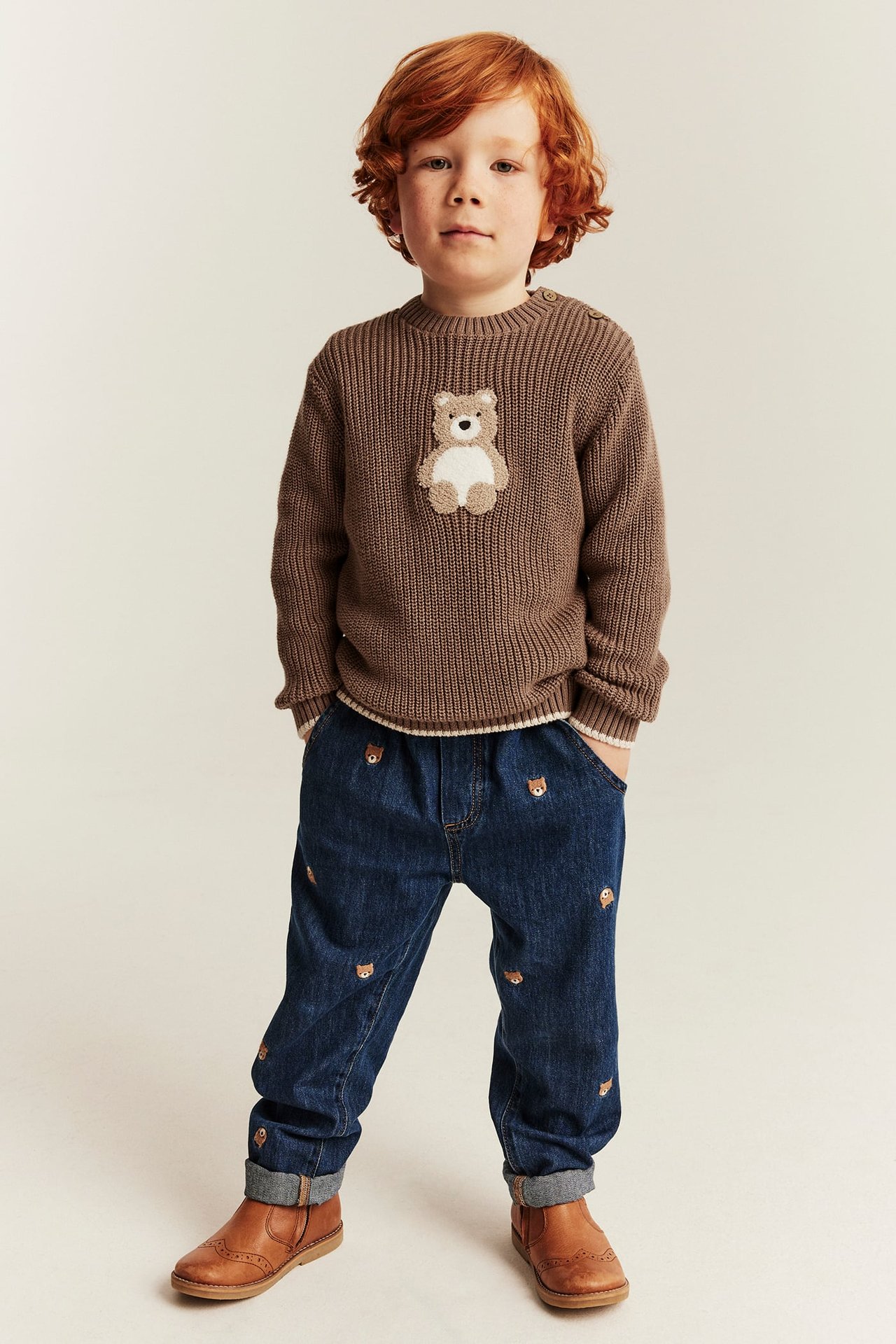 Barn i brun ribbestrikket teddybjørn-genser, mørkeblå rev-broderte jeans og brune skinnstøvler.