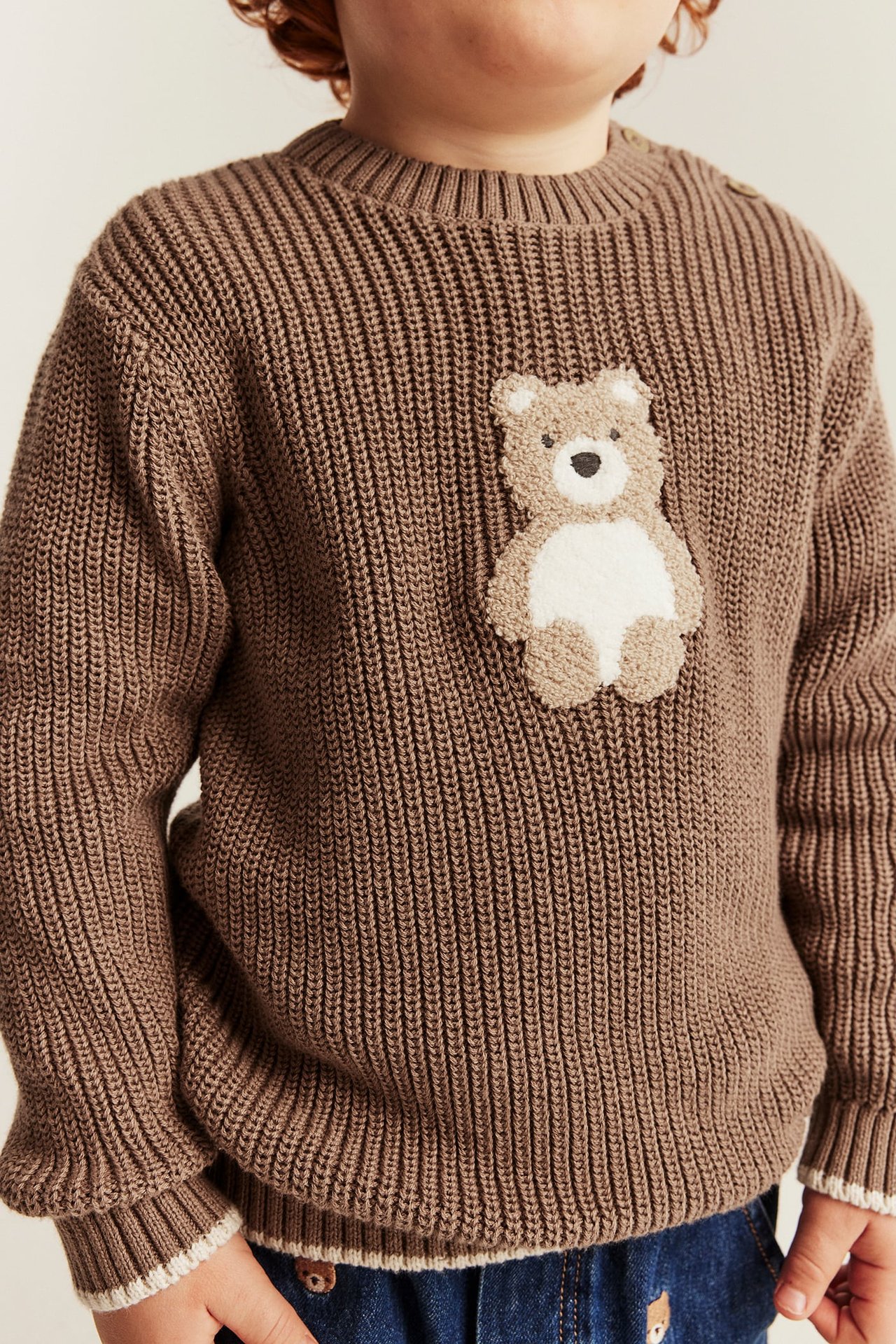 Frontutsikt, barns brun ribbestrikket genser med teddybjørnapplikasjon og blå jeans.