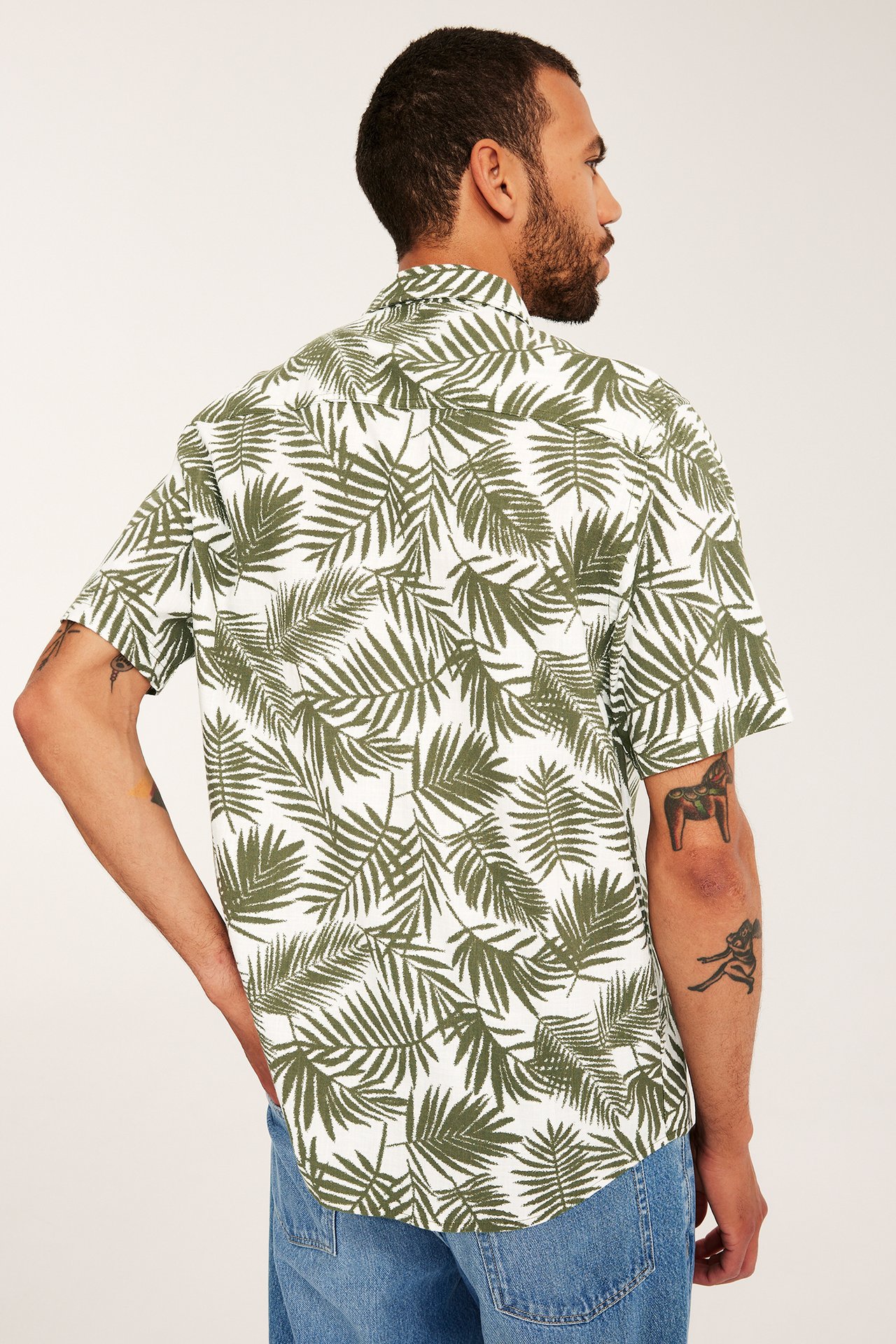 Bakifrån/sida vy av man i vit och khaki-grön palmbladsmönstrad kortärmad skjorta och blå jeans.