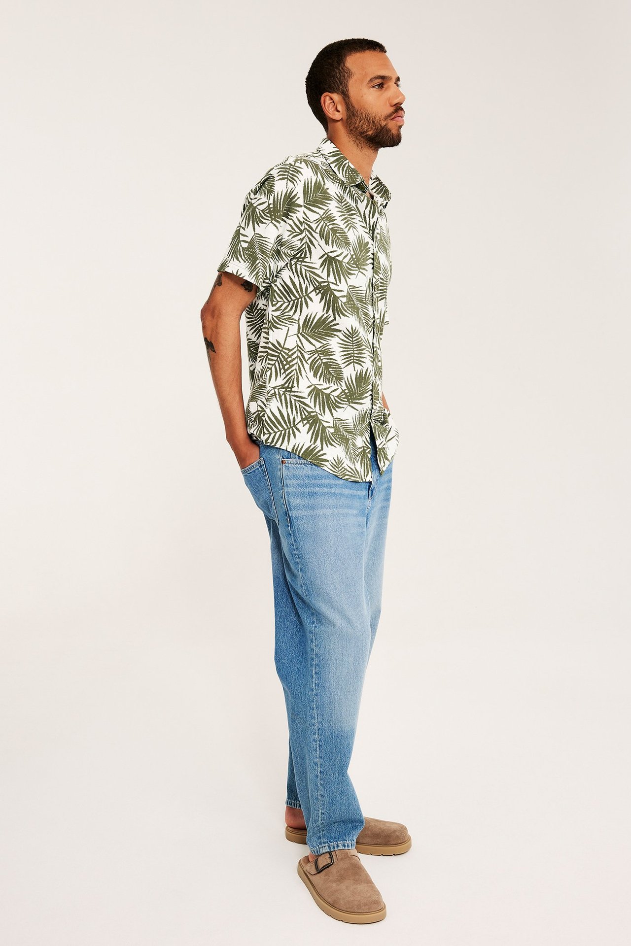 Sidovy: man i en vit och khakigrön palmbladmönstrad kortärmad skjorta, ljusblå jeans och bruna mockasandaler.