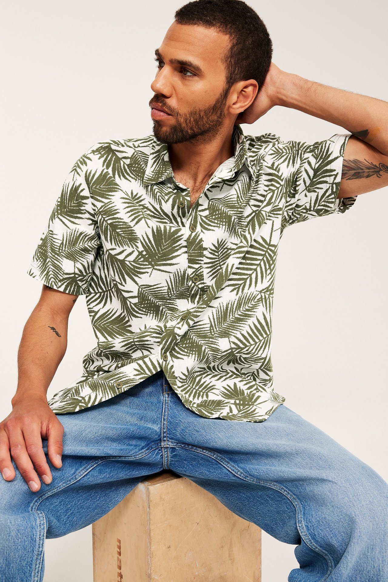 Framifrån, en man i en vit och khakigrön palmbladmönstrad kortärmad bomullsskjorta och ljusblå jeans.