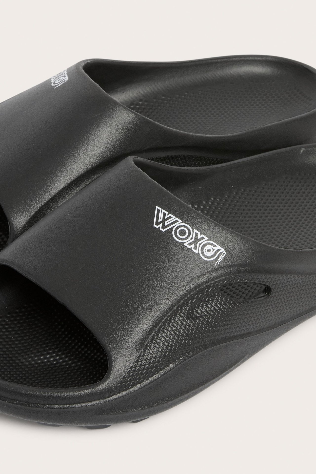Närbild av svarta plastsandaler med vit WOXO-logga på remmen och texturerade sulor.