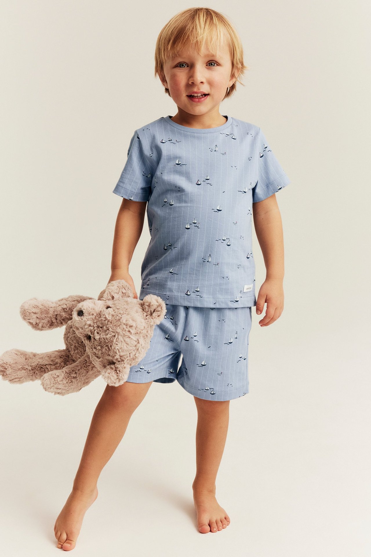 Lapsi vaaleansinisessä puuvillapyjamasetissä, lyhyet hihat ja venekuvio. Pitää ruskeaa nallekarhua.