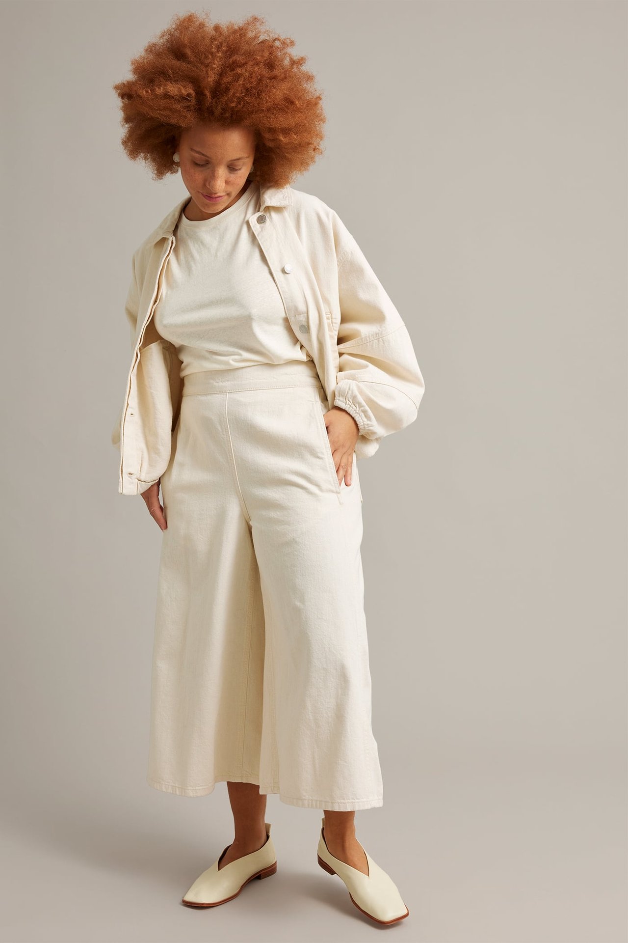 Frontal: kvinne i beige bomulls culottejeans, matchende topp, jakke og lyse flate sko.