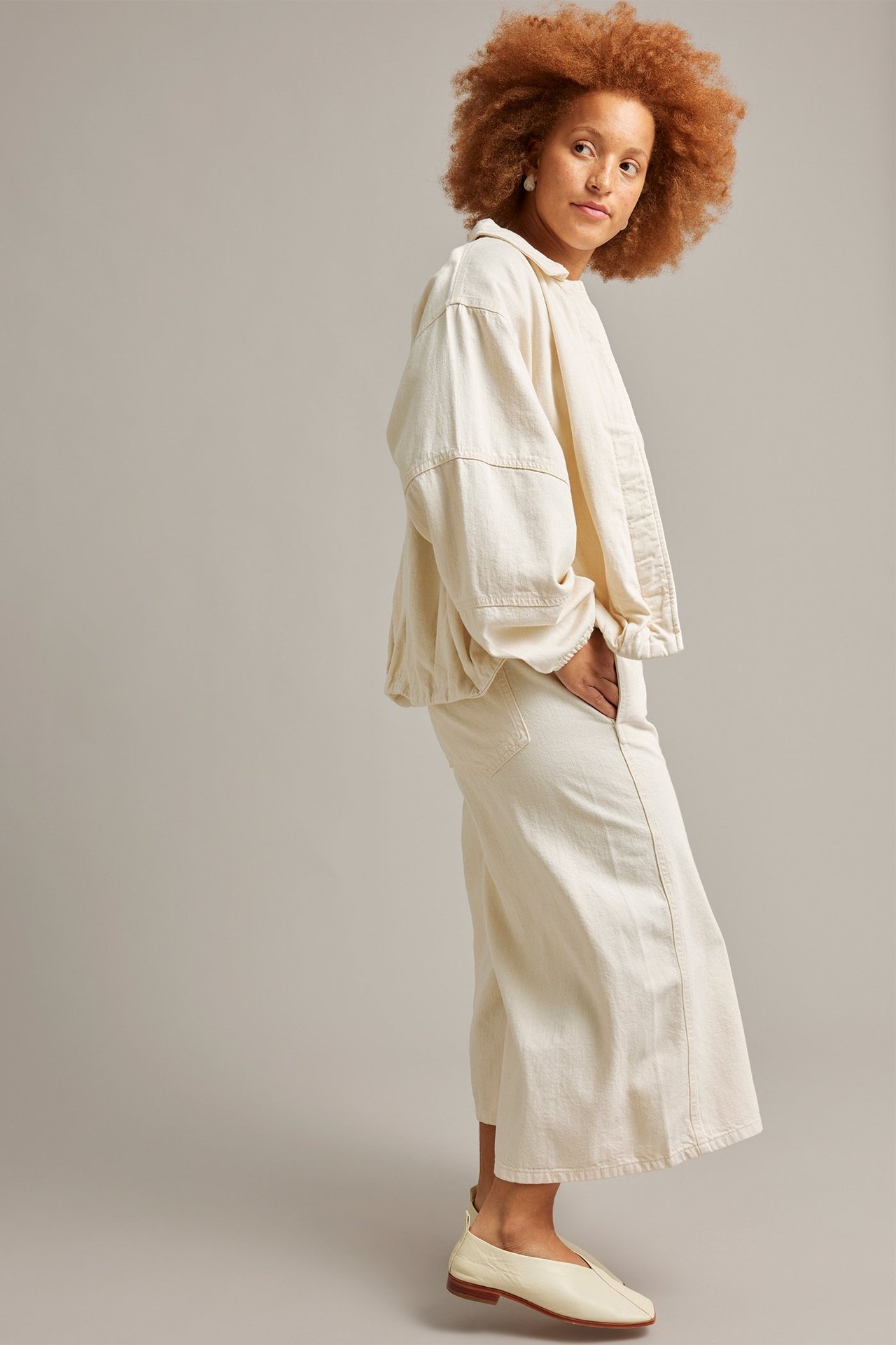 Sidebilde: Kvinne i beige, cropped pull-on culottejeans, matchende denimjakke og beige flate sko.