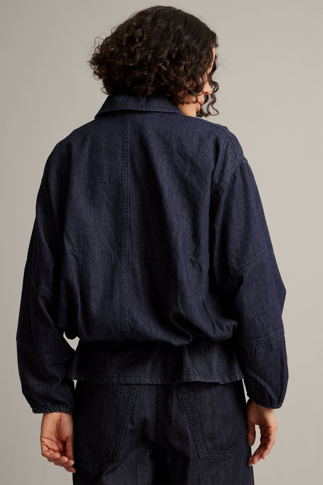 Bakifrån: kvinna i mörkblå denimjacka med muddar och matchande byxor.