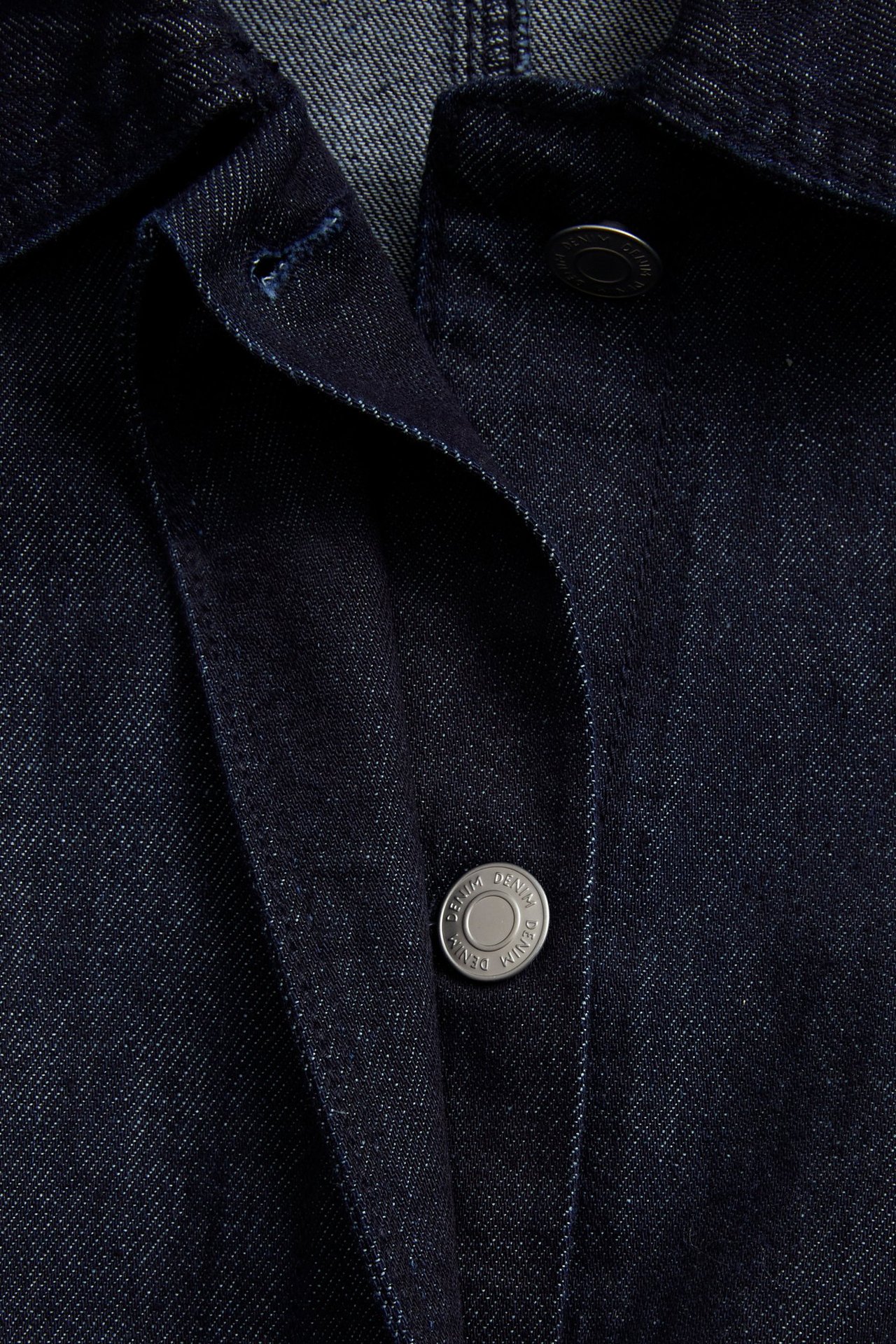 Lähikuva edestä tummasta denim-puserojakista, jossa on hopeinen metallinappi.