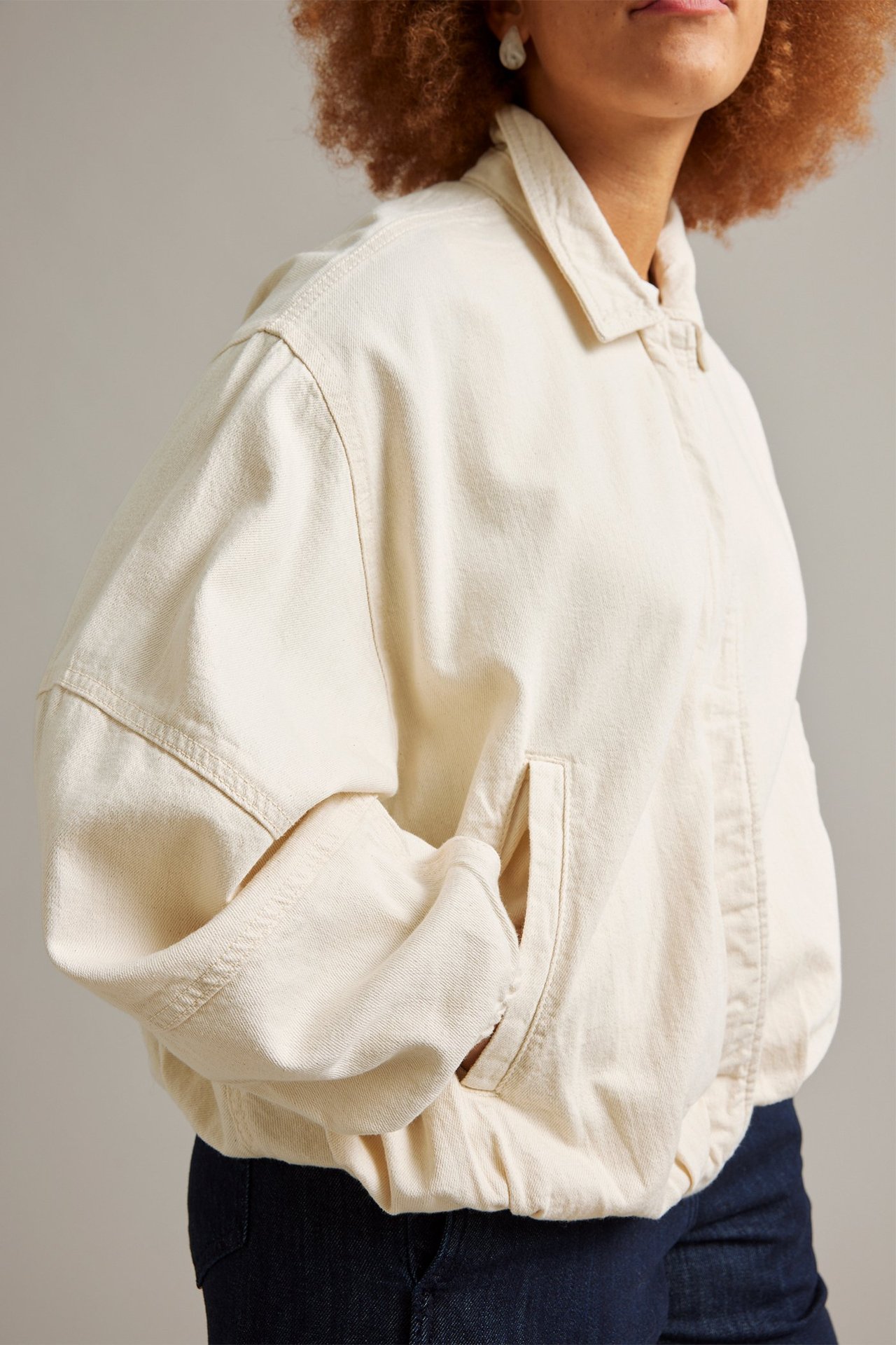 Sivukuva naisesta beigessä puuvilladenim bomber-takissa, jossa pystytaskut, ja tummansinisissä farkuissa.