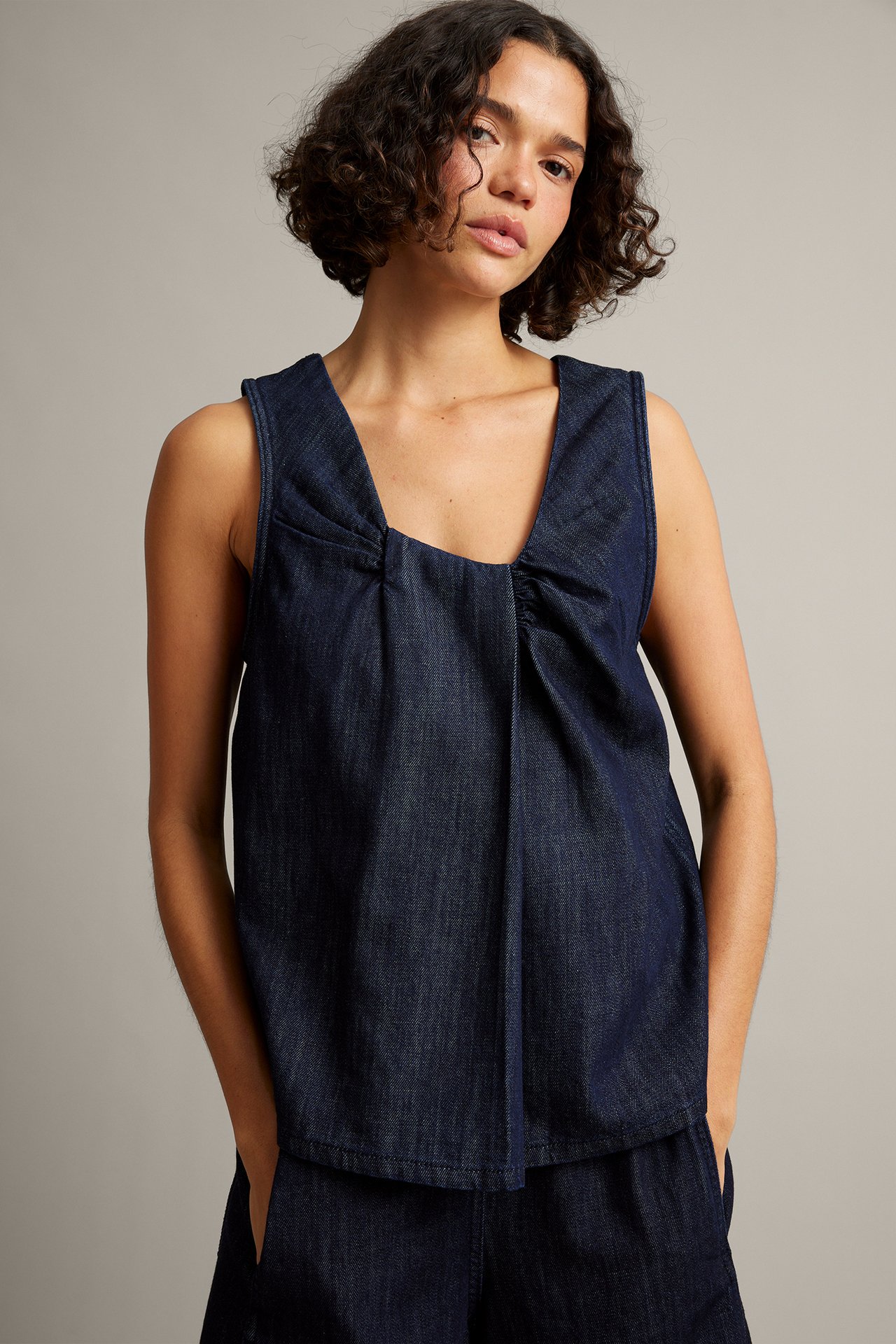Frontvy, kvinnlig modell i mörkblå ärmlös topp i denim med veckad halslinning och matchande shorts.