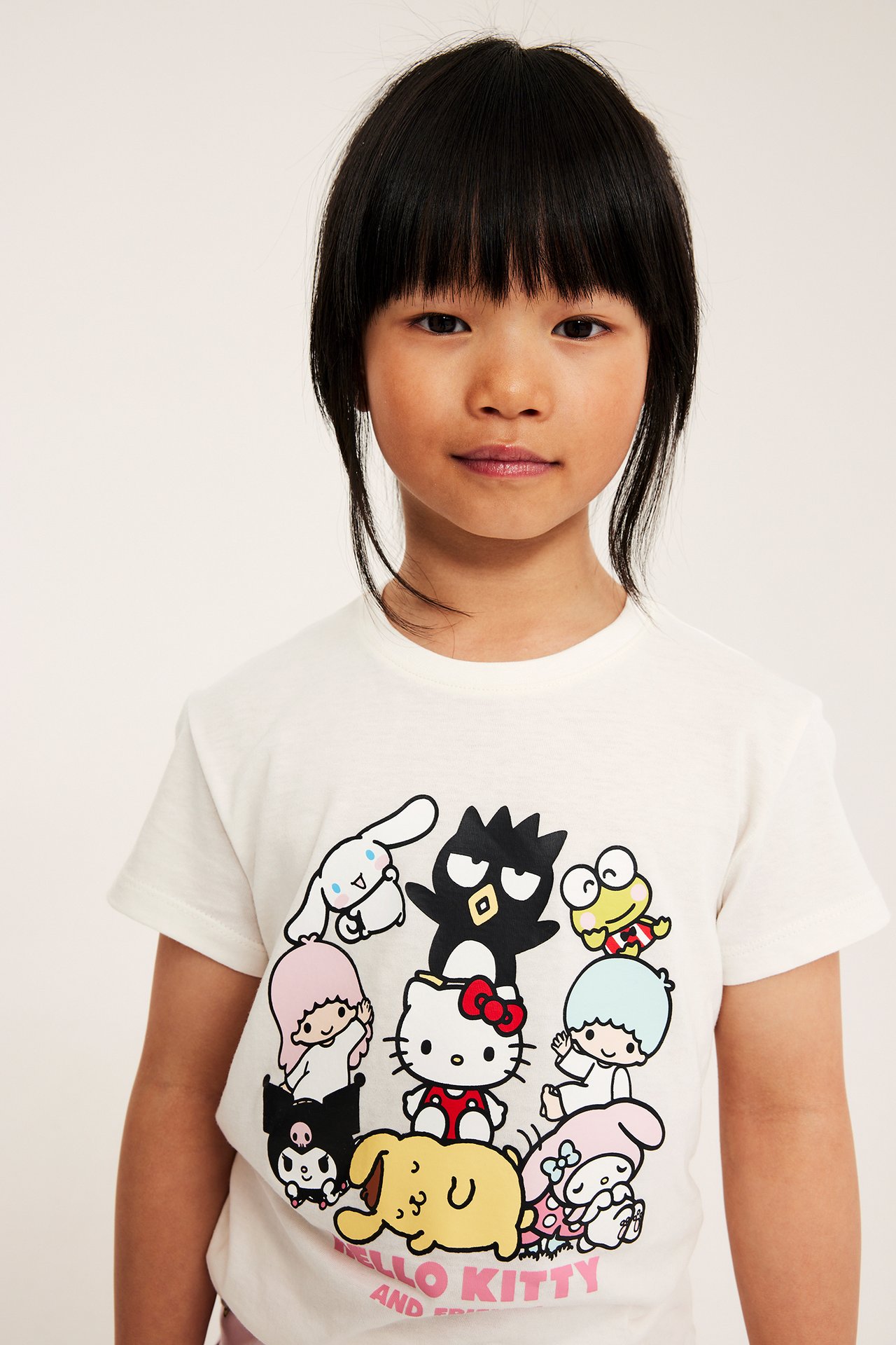 Edestä näkymä lapsesta, joka käyttää luonnonvalkoista lyhythihaista T-paitaa Hello Kitty ja ystävät -printillä.