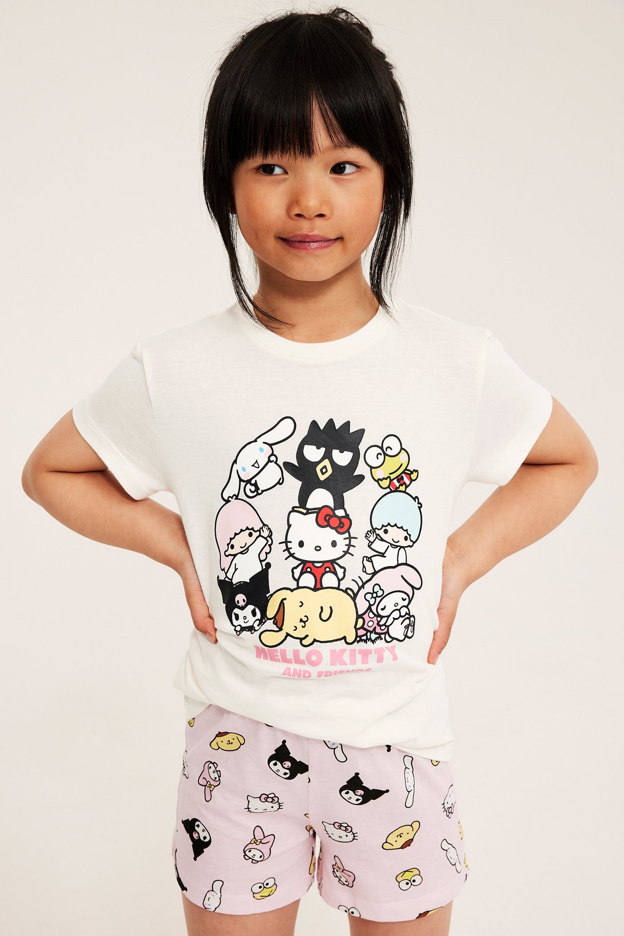Lapsi pukeutuu luonnonvalkoiseen Hello Kitty -toppiin ja vaaleanpunaisiin kuviollisiin shortseihin. Edestä.