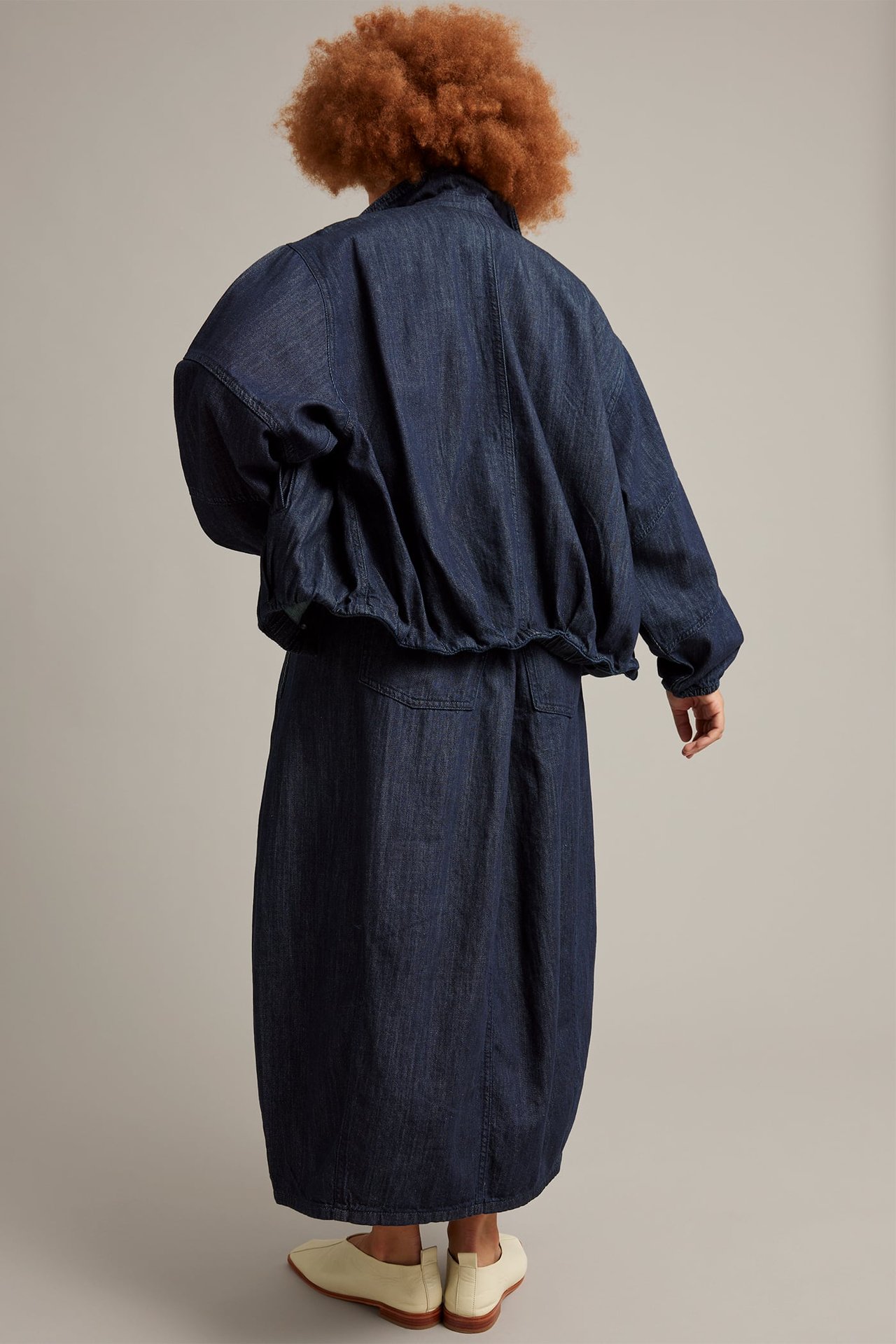 Takaa: nainen tummansinisessä barrel-denimhameessa taskuilla, yhteensopivassa bomber-takissa ja vaaleanbeigessä tasakengissä.