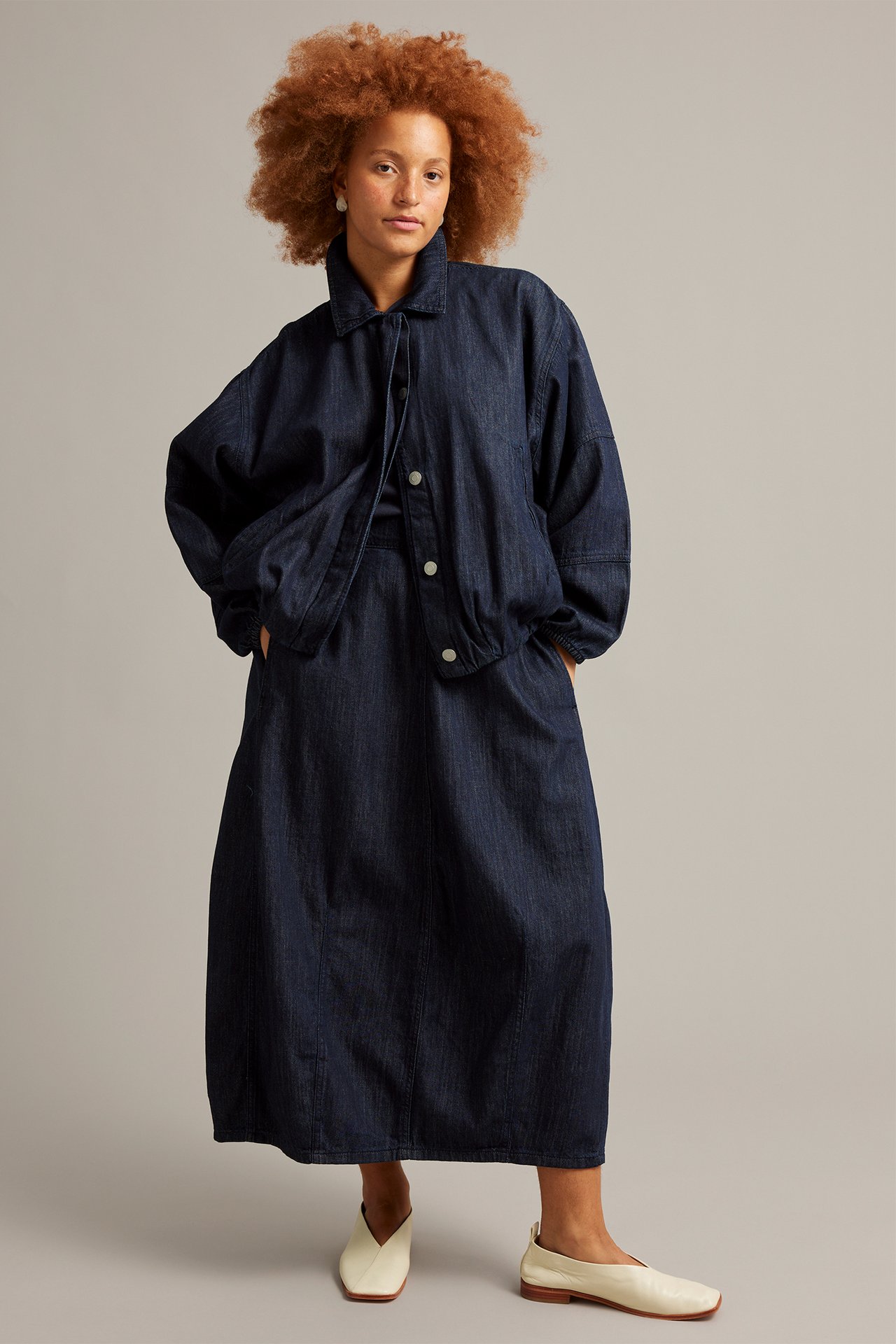Edestäpäin nainen tummassa denim-barrelhameessa ja yhteensopivassa takissa kermanvärisillä kengillä.