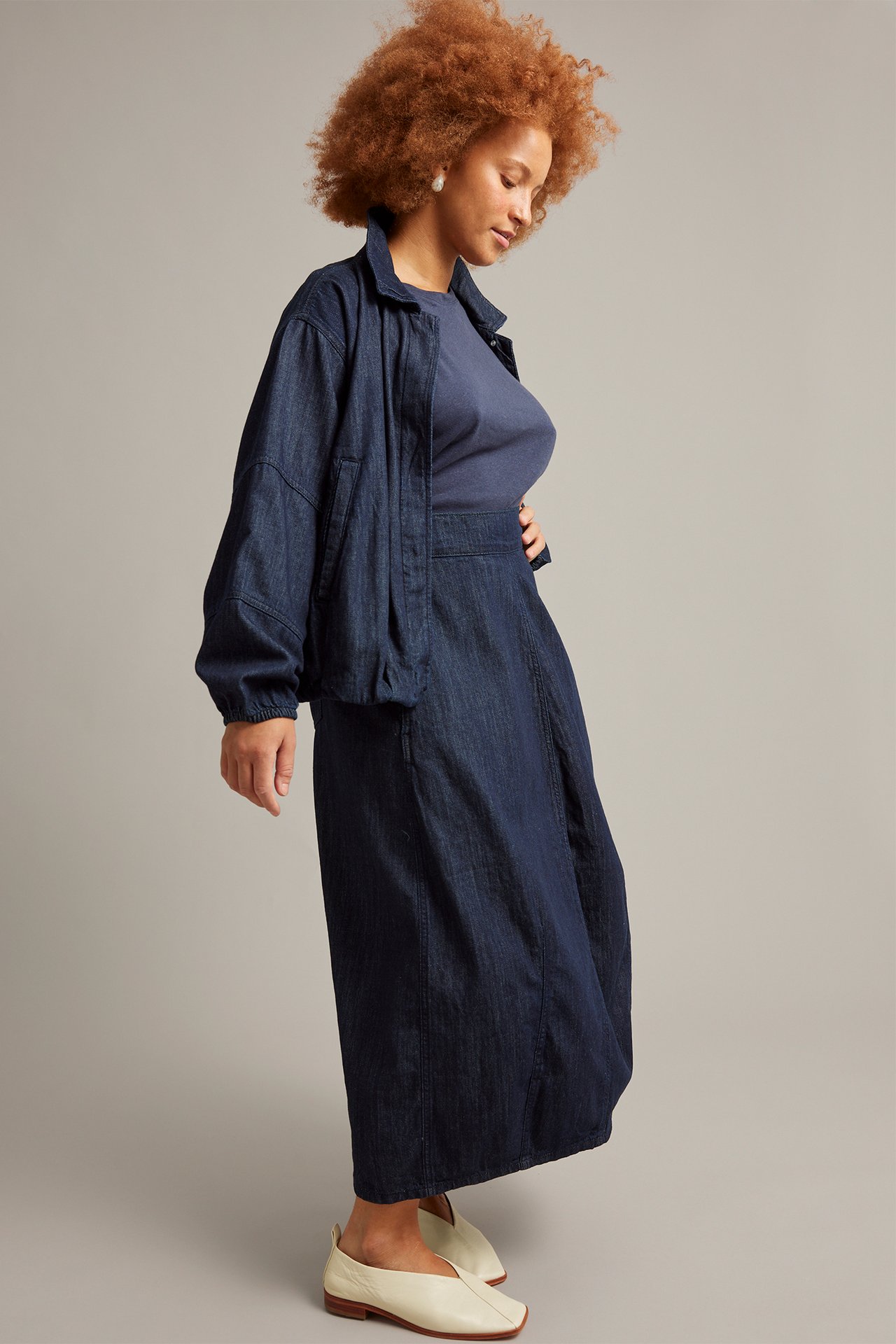 Naisen sivukuva tummansinisessä denim barrel-hameessa, tummassa denimtakissa, laivastonsinisessä t-paidassa ja kermanvärisissä kengissä.