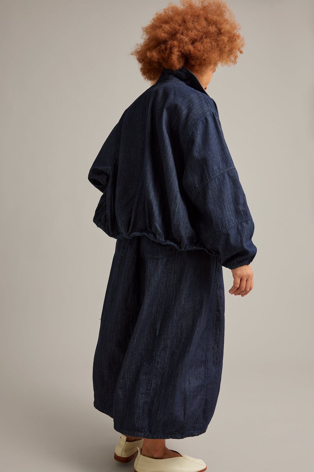 Takaa ja sivulta: Nainen tummansinisessä barrel-denimhameessa taskuilla, denimtakissa ja vaaleissa kengissä.
