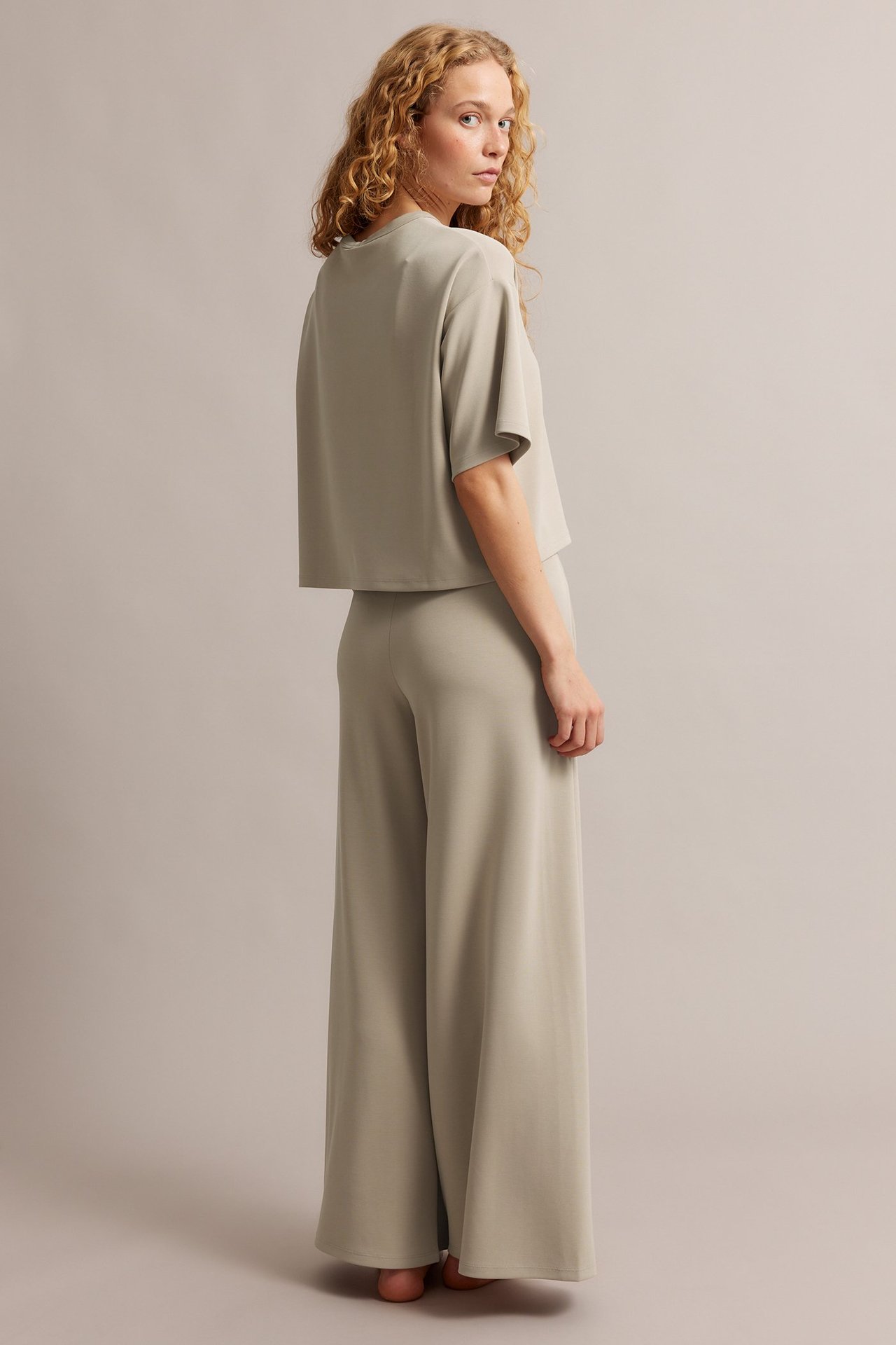Kvinna i bakifrån-sidovy, bärandes beiga raka loungebyxor och en matchande kortärmad topp.
