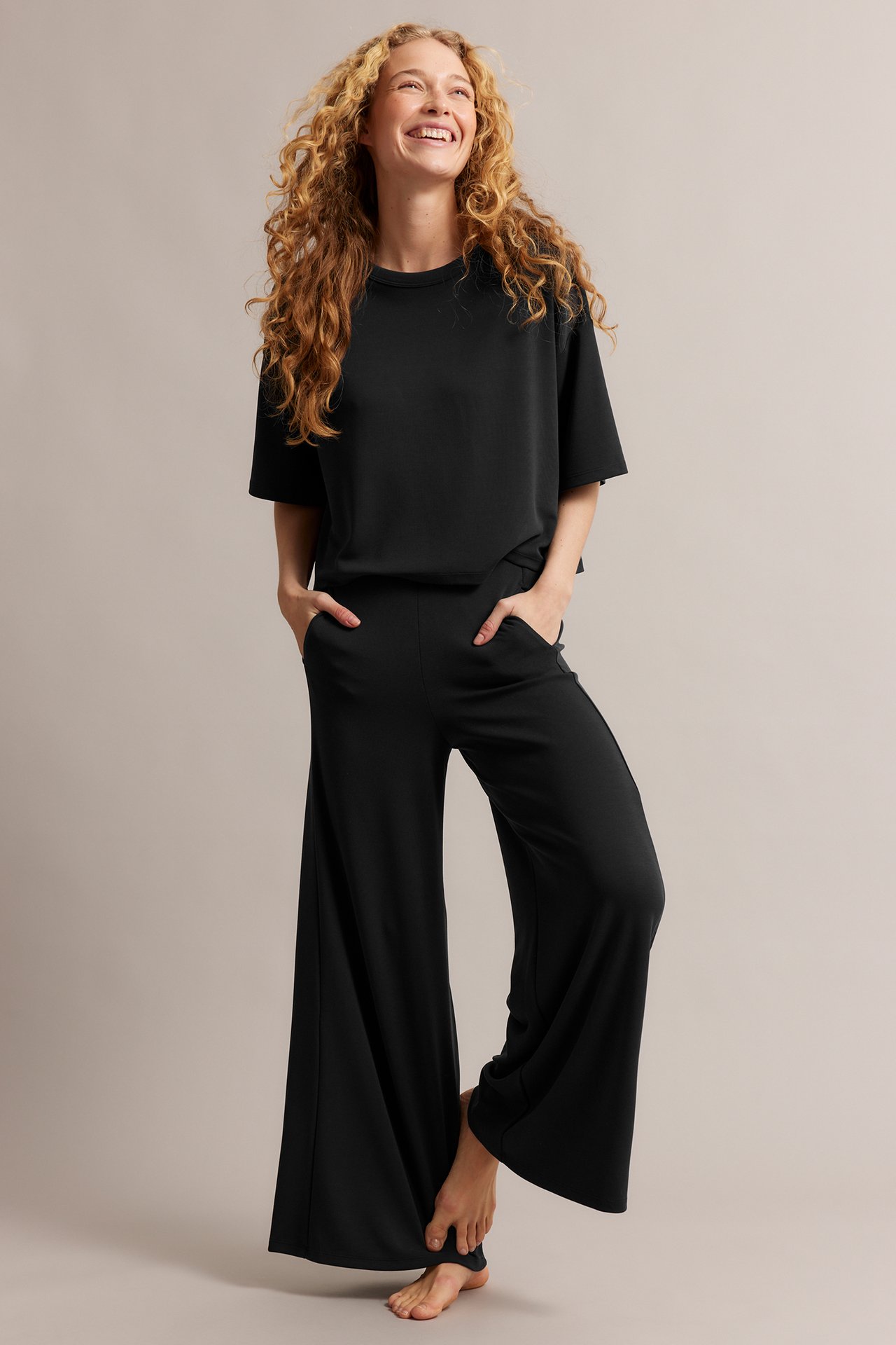 Hymyilevä nainen edestä, yllään musta laatikkomainen T-paita ja mustat leveälahkeiset housut, kädet taskuissa.