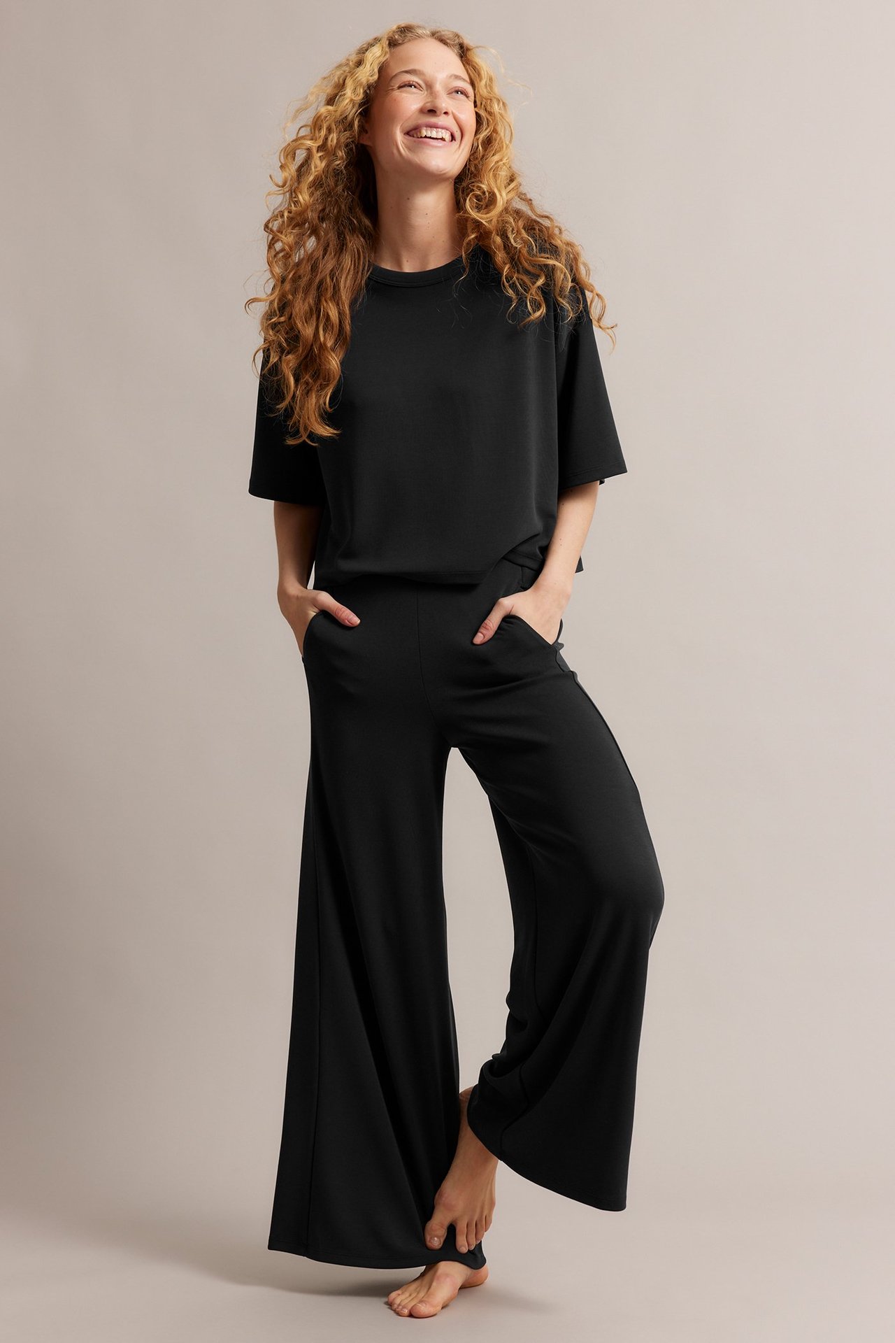 Hymyilevä nainen edestä, yllään musta laatikkomainen T-paita ja mustat leveälahkeiset housut, kädet taskuissa.