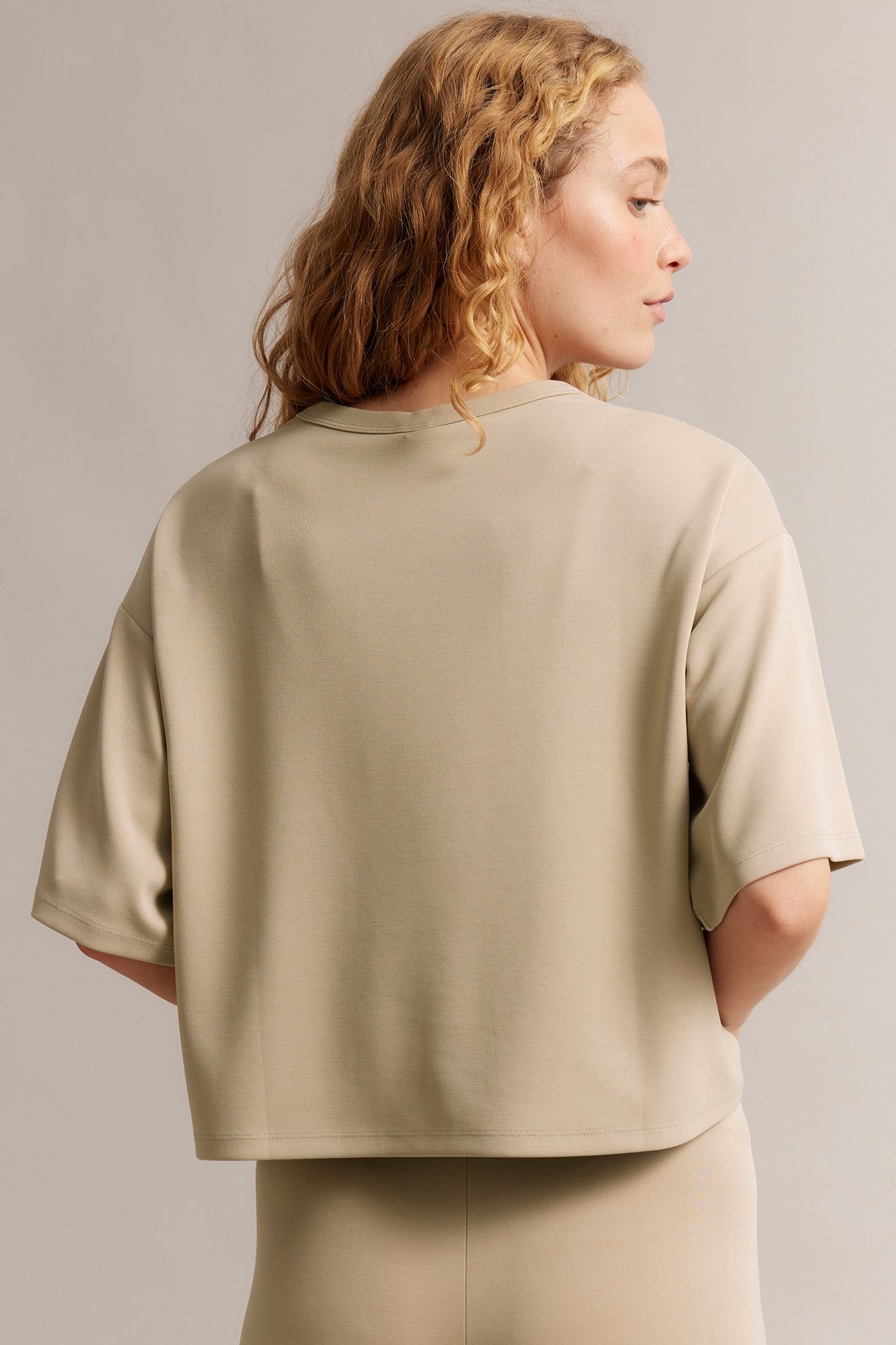 Nainen takaa: beige laatikkomainen T-paita pidemmillä hihoilla ja yhteensopivat beiget alaosat.