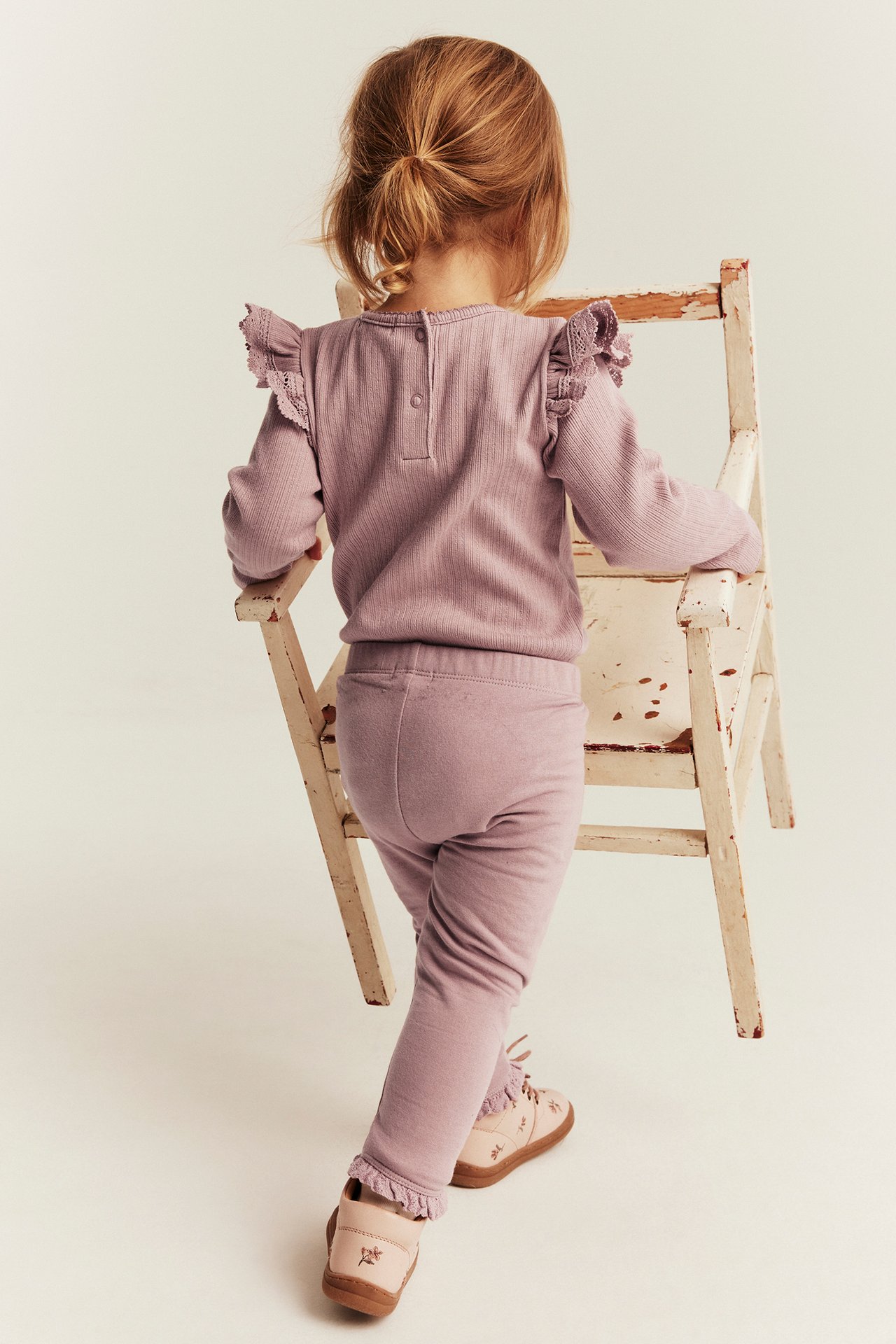 Bakifrån: barn i lila ribbad långärmad tröja med spets, matchande leggings och broderade skor.