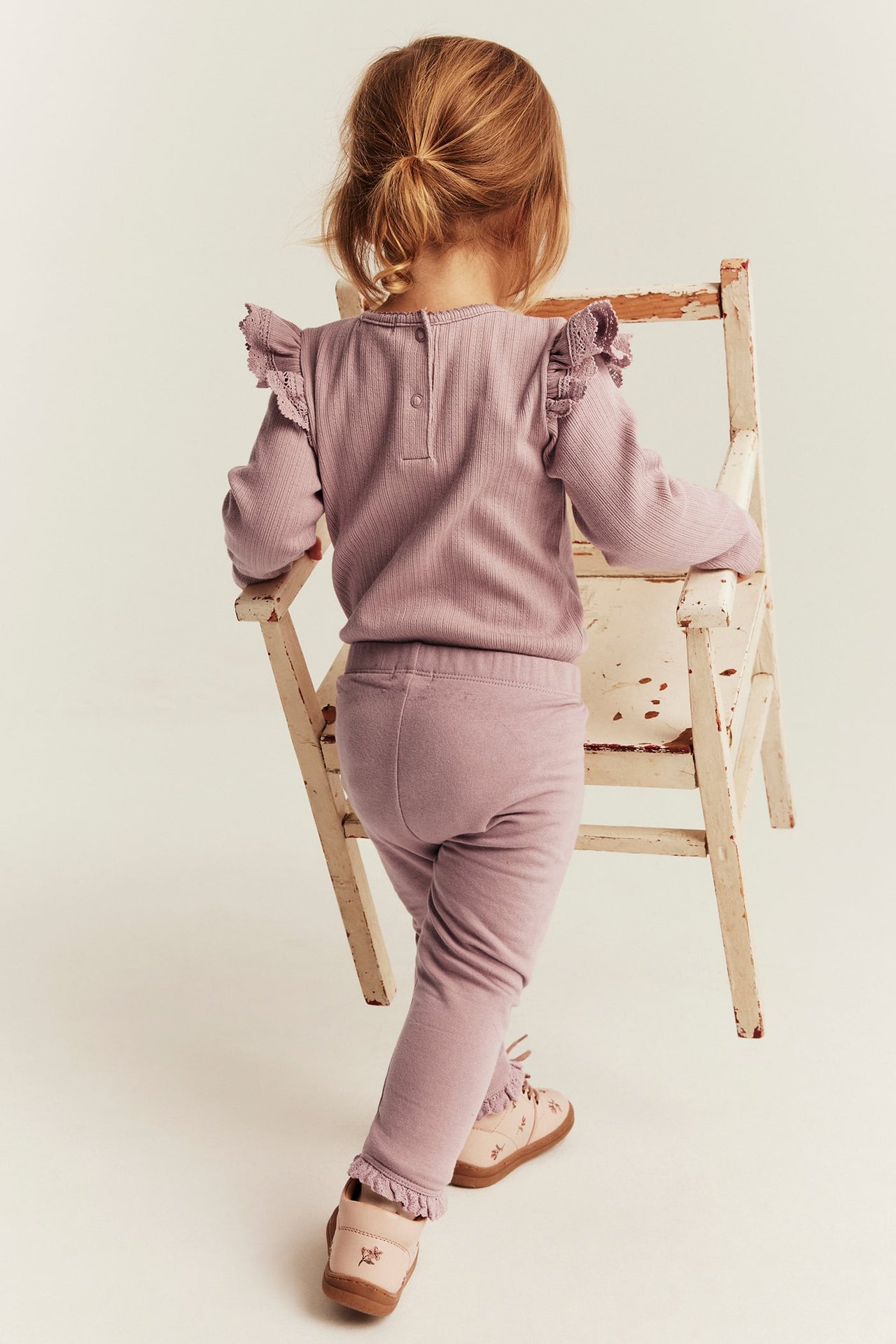 Bakifrån: barn i lila ribbad långärmad tröja med spets, matchande leggings och broderade skor.