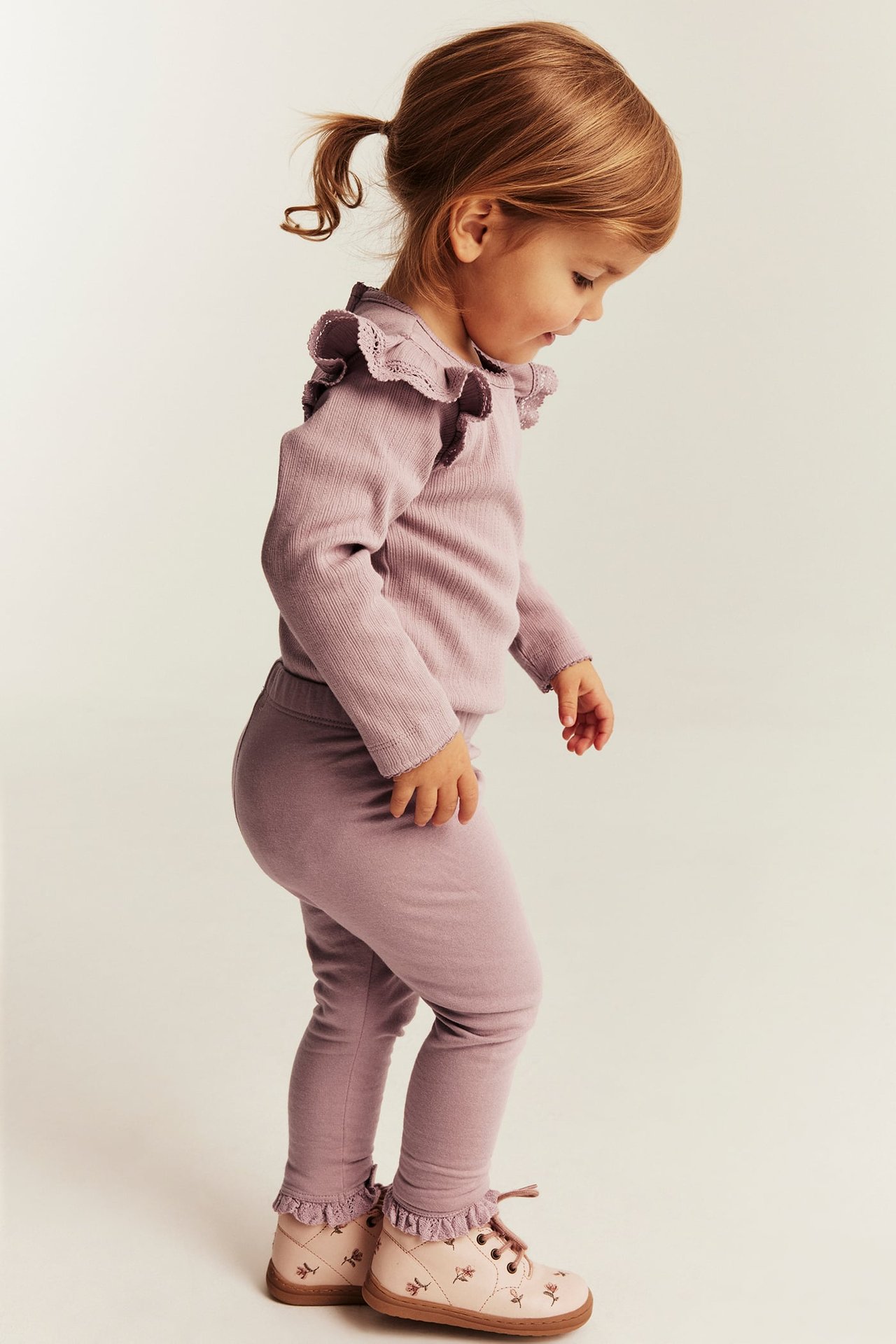 Ett barn bär en dammlila ribbad topp med volanger, matchande leggings och rosa blommiga skor.