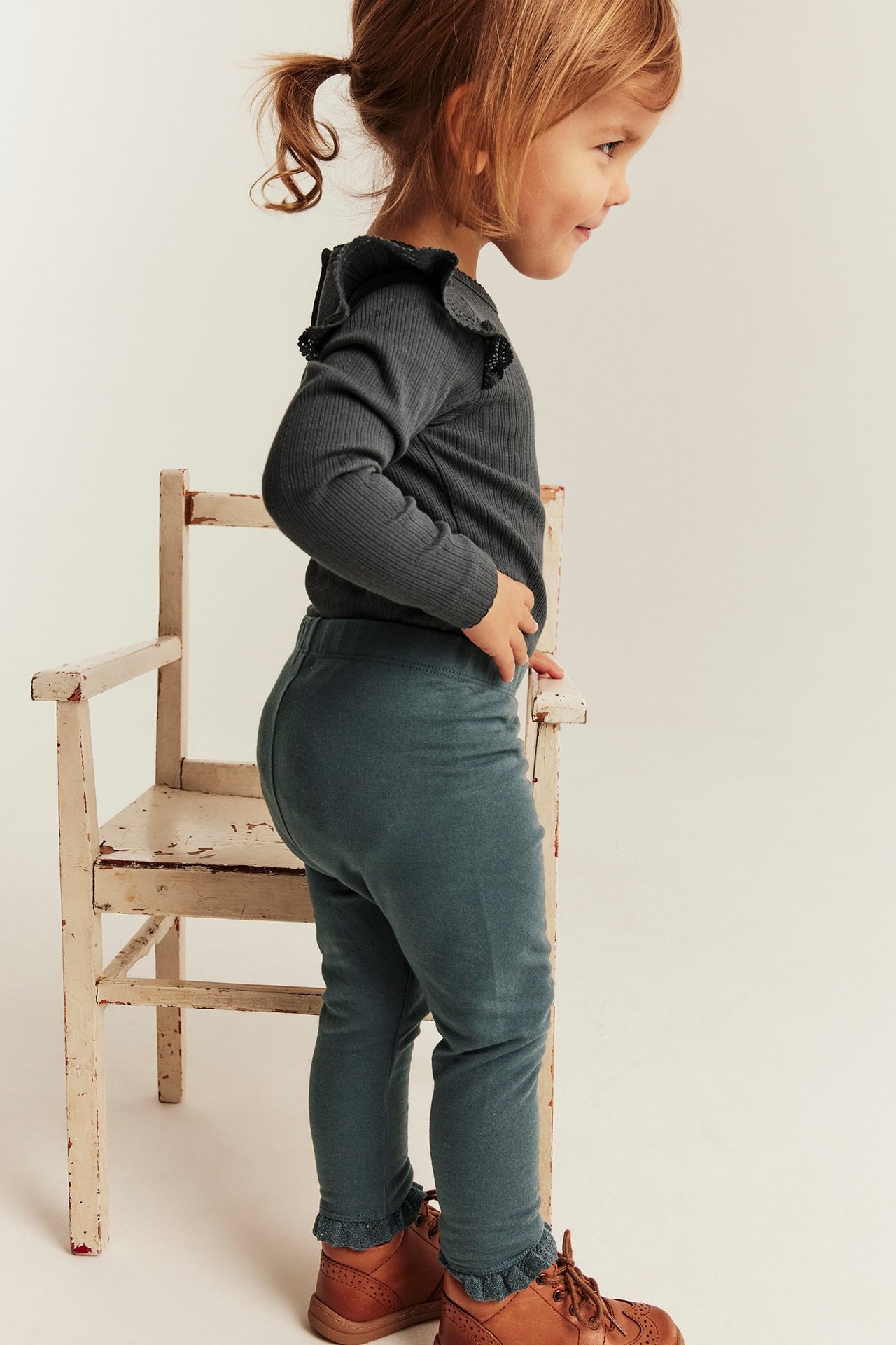Sidestående barn i teal leggings, mørkegrå ribbestrikket topp med rysjer og brune støvler.