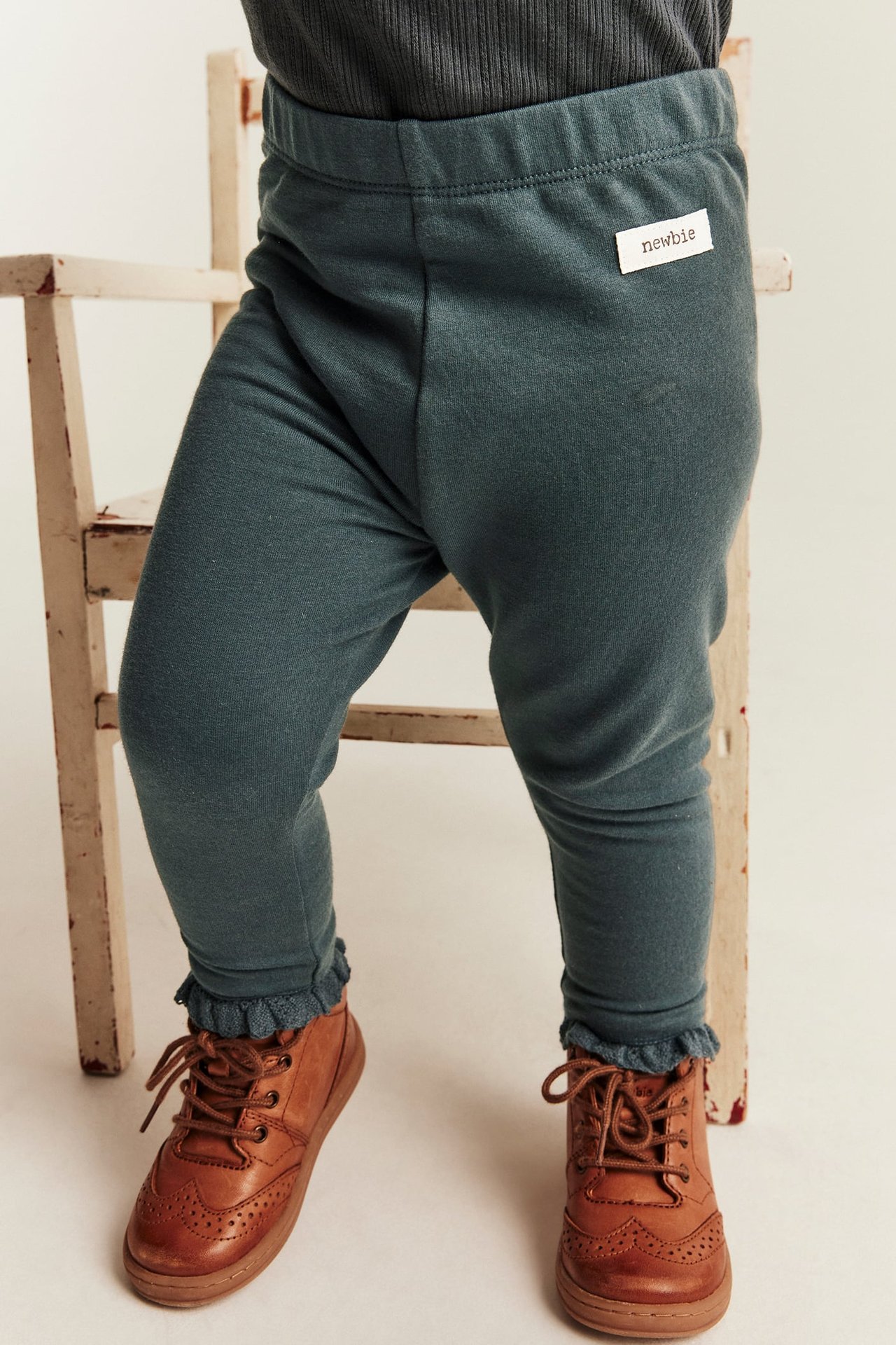 Baby i mørk teal jerseyleggings med rysjekanter, mørk grå topp og brune snørestøvler.