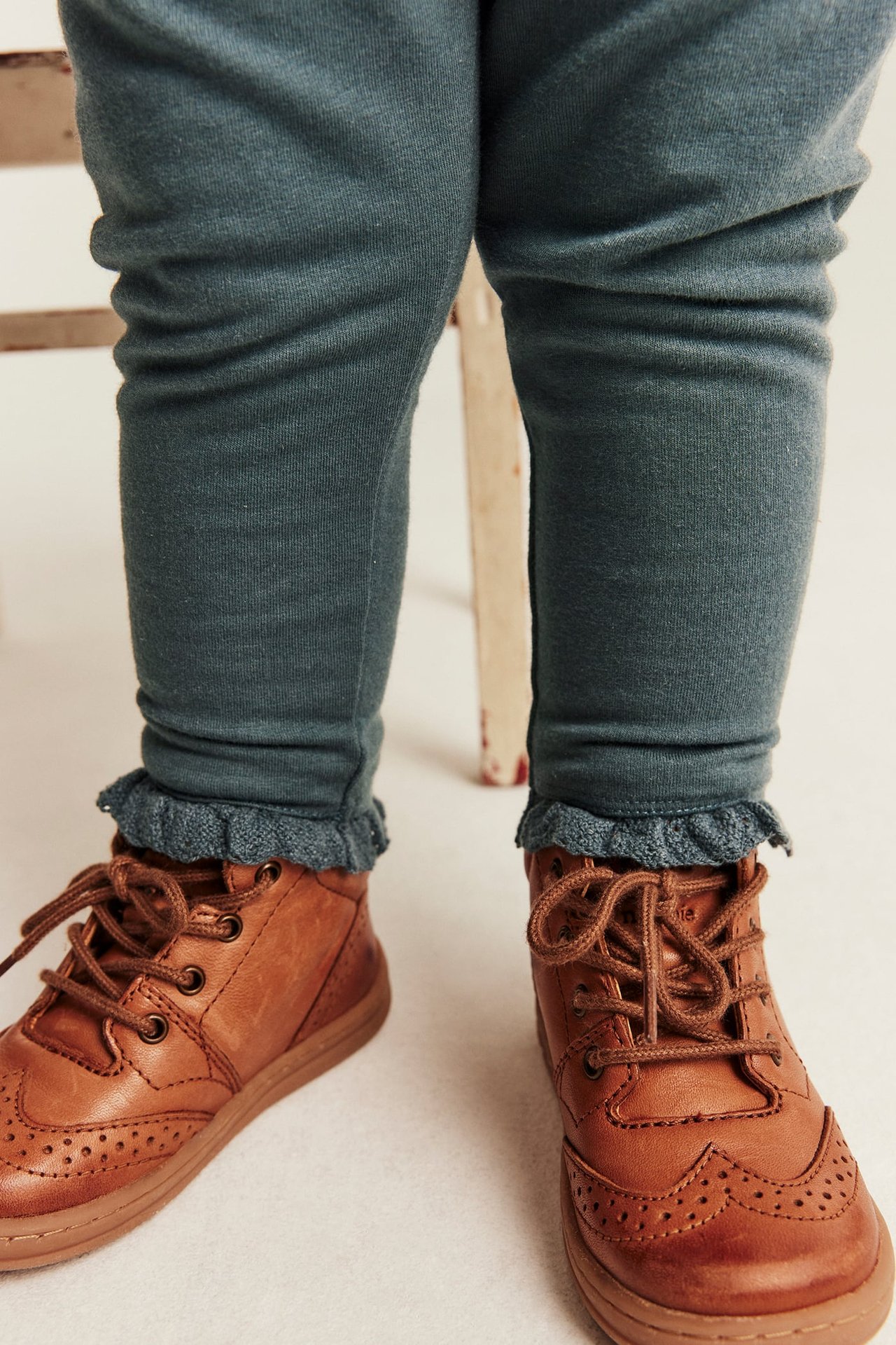 Nærbilde forfra av barnets mørkegrønne jerseyleggings med rysjekanter over brune snørestøvler.