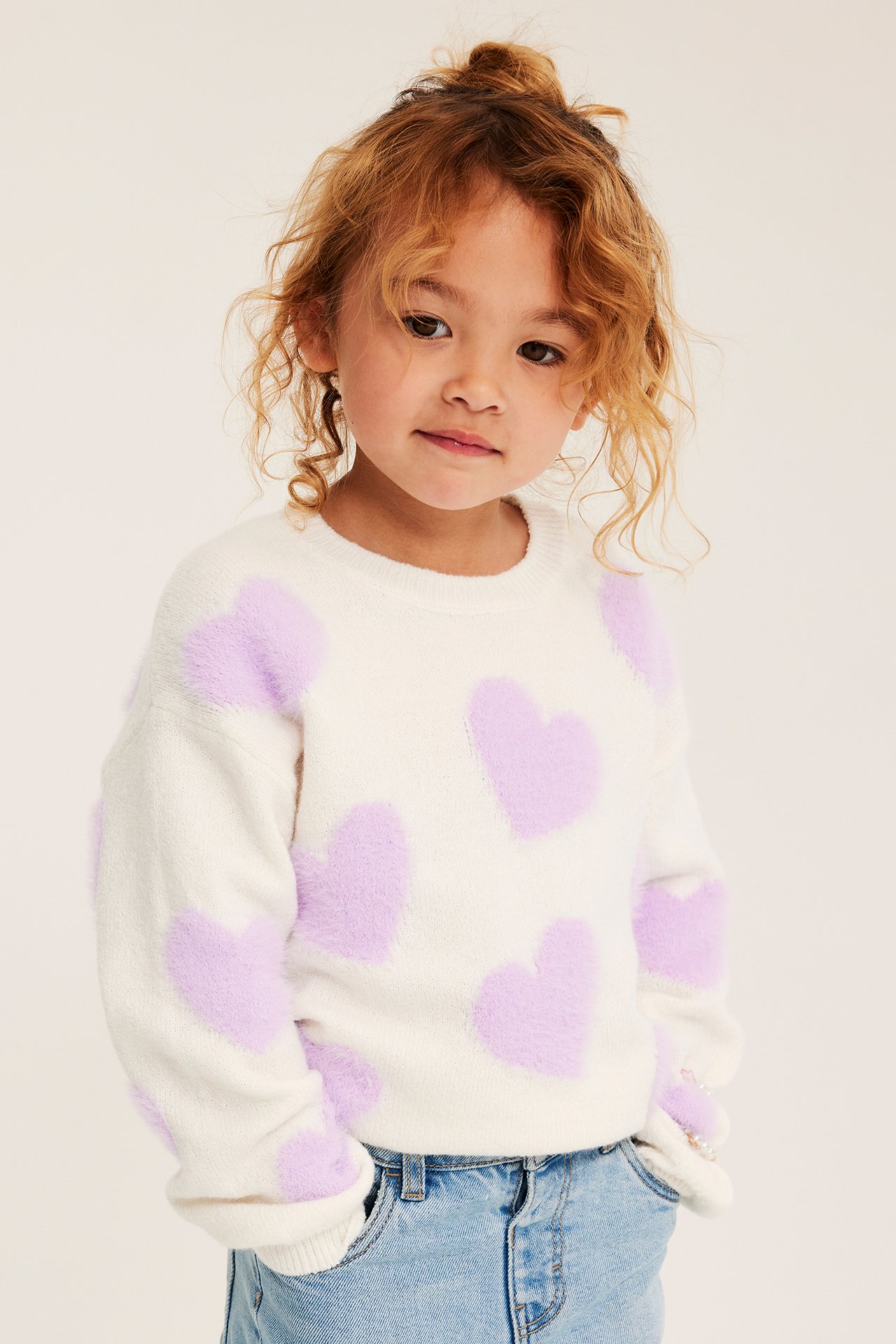 Frontvy av ett barn i vit fluffig tröja med lila hjärtan och blå jeans.