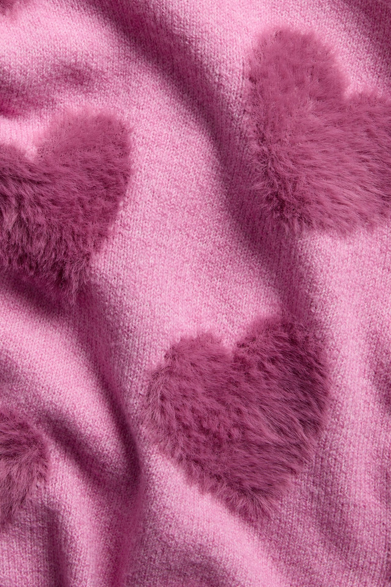 Närbild på ett rosa stickat tyg med mörkrosa lurviga hjärtapplikationer.
