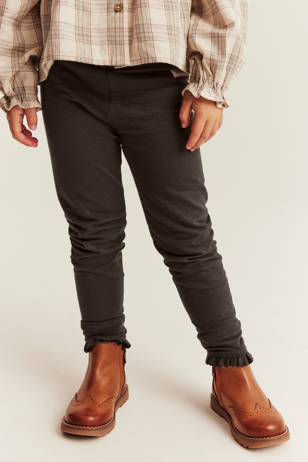 Edestäpäin lapsi tummanharmaissa leggingseissä, joissa röyhelöreunat, beige ruutupaita ja ruskeat broguekengät.