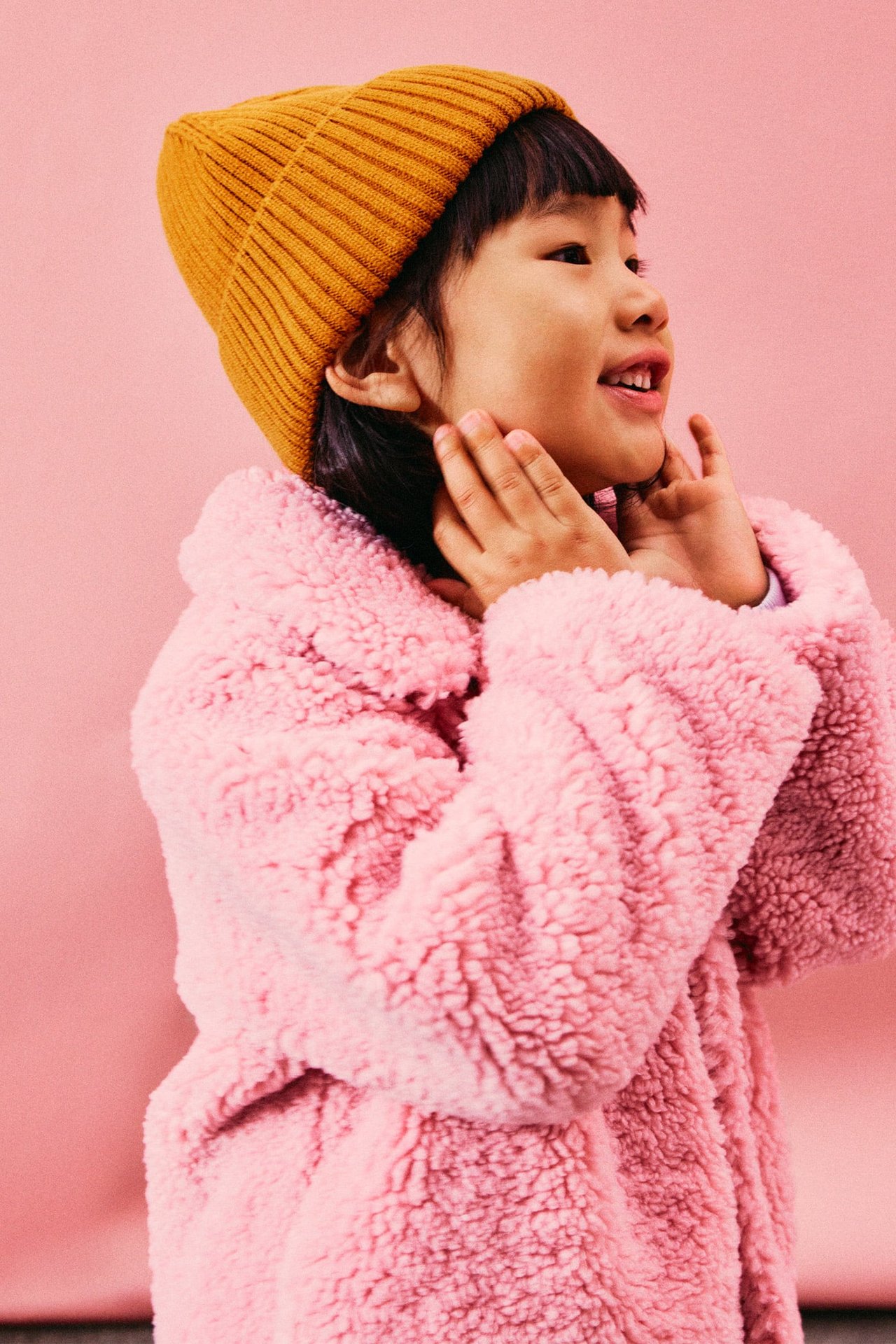 Kvinna i rosa fluffig jacka och orange stickad mössa, ståendes i profil mot rosa bakgrund