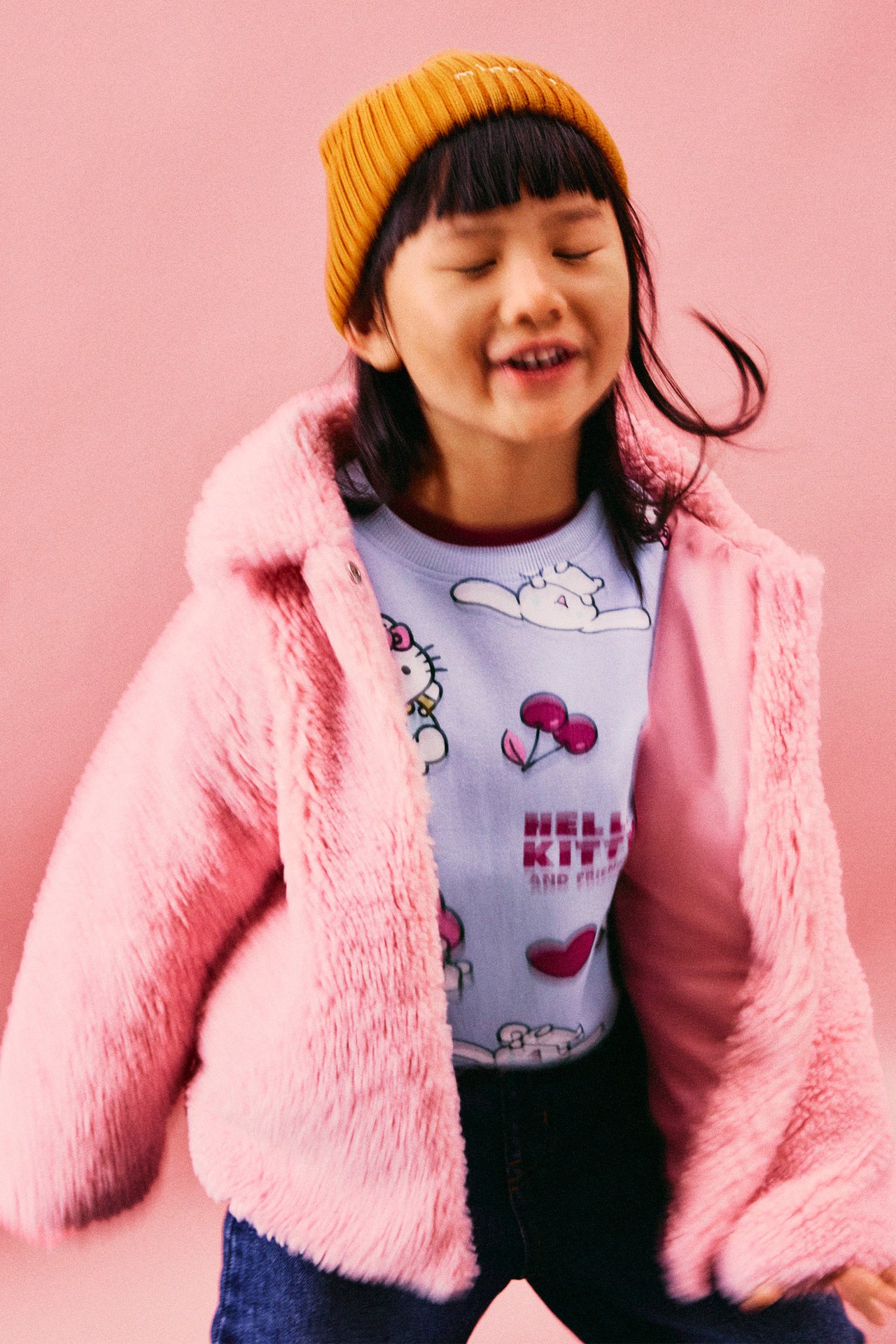 Barn med svart hår, stående framifrån, bär ljusblå t-shirt med tryck och rosa fluffig jacka med huva