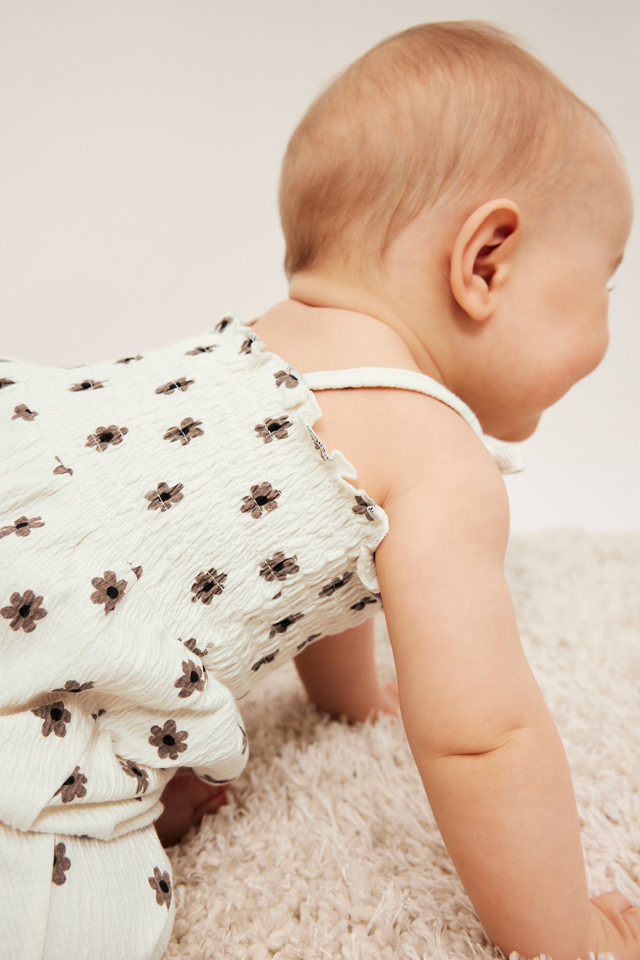 Side-bakfra: baby i offwhite blomstret krøllet jumpsuit med smocket topp og sløyfer.