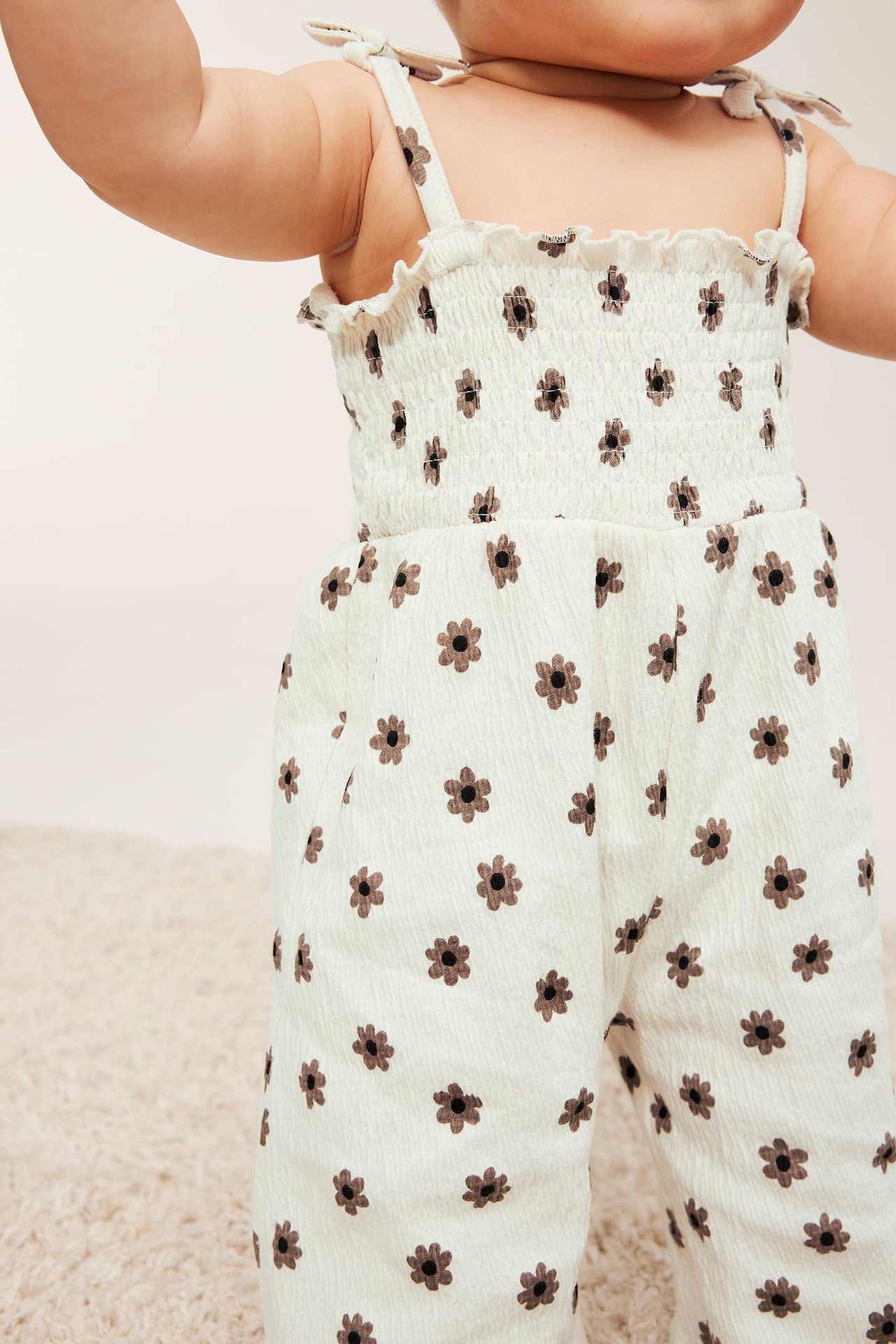 Frontvy av en baby i en offwhite blommig jumpsuit med smockad topp och rosettband.