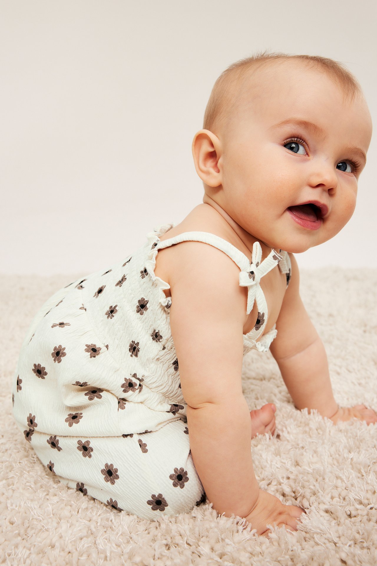 Sidevisning av en baby i en offwhite blomstret krøllete jumpsuit med smocked overdel og sløyfebånd.