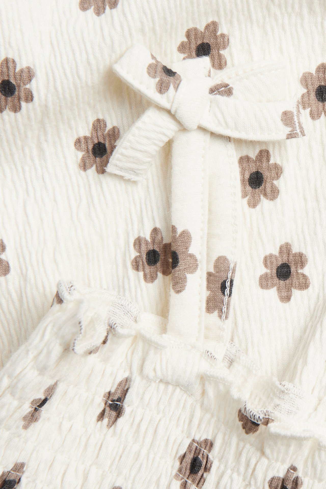 Edestä lähikuva: Luonnonvalkoinen ryppyinen vauvanhaalari ruskealla kukkakuviolla, smokkiryppy ja olkainrusetti.