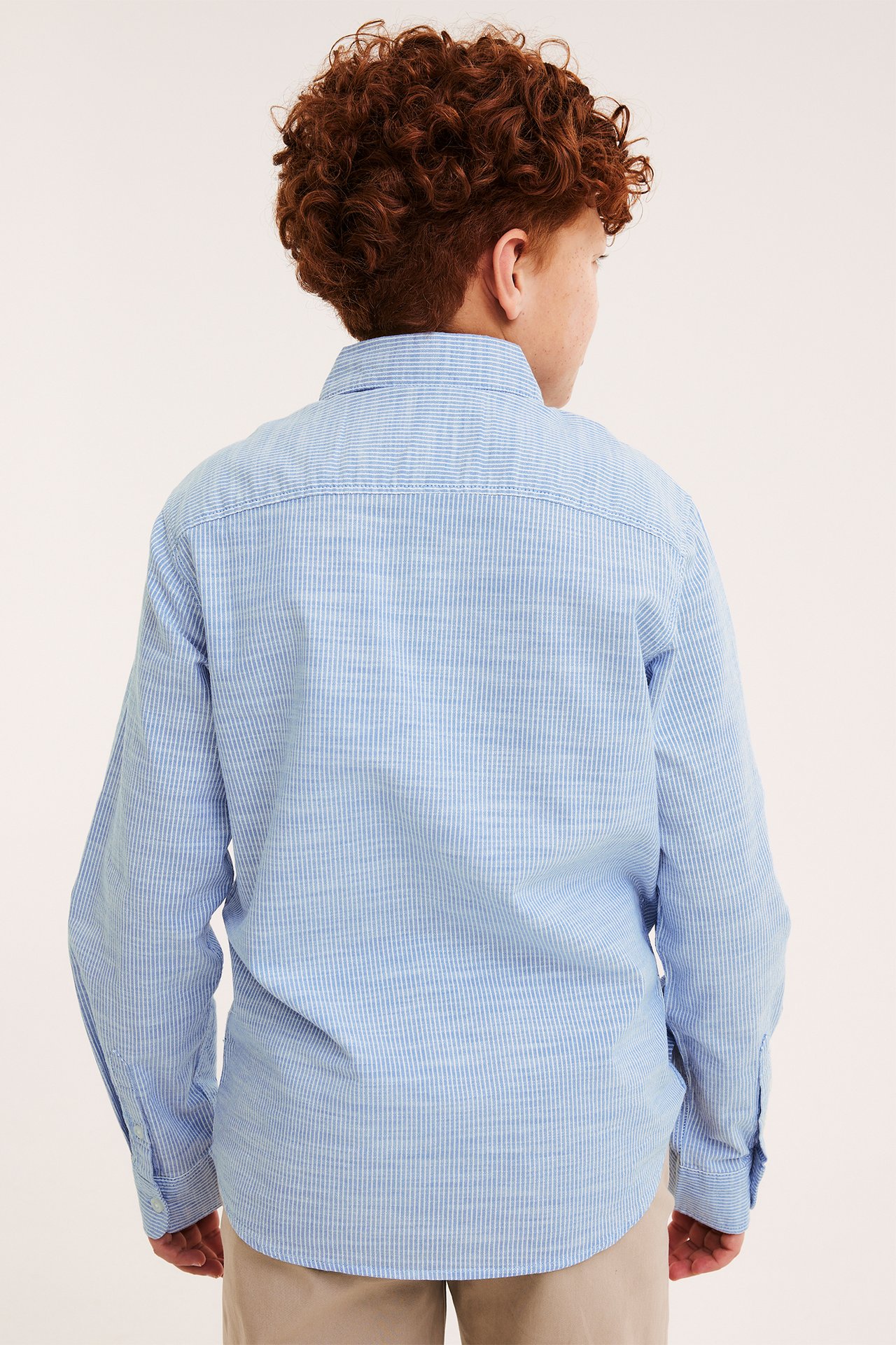 Bakifrån, ett barn i en ljusblå randig långärmad bomullsskjorta och beige byxor.