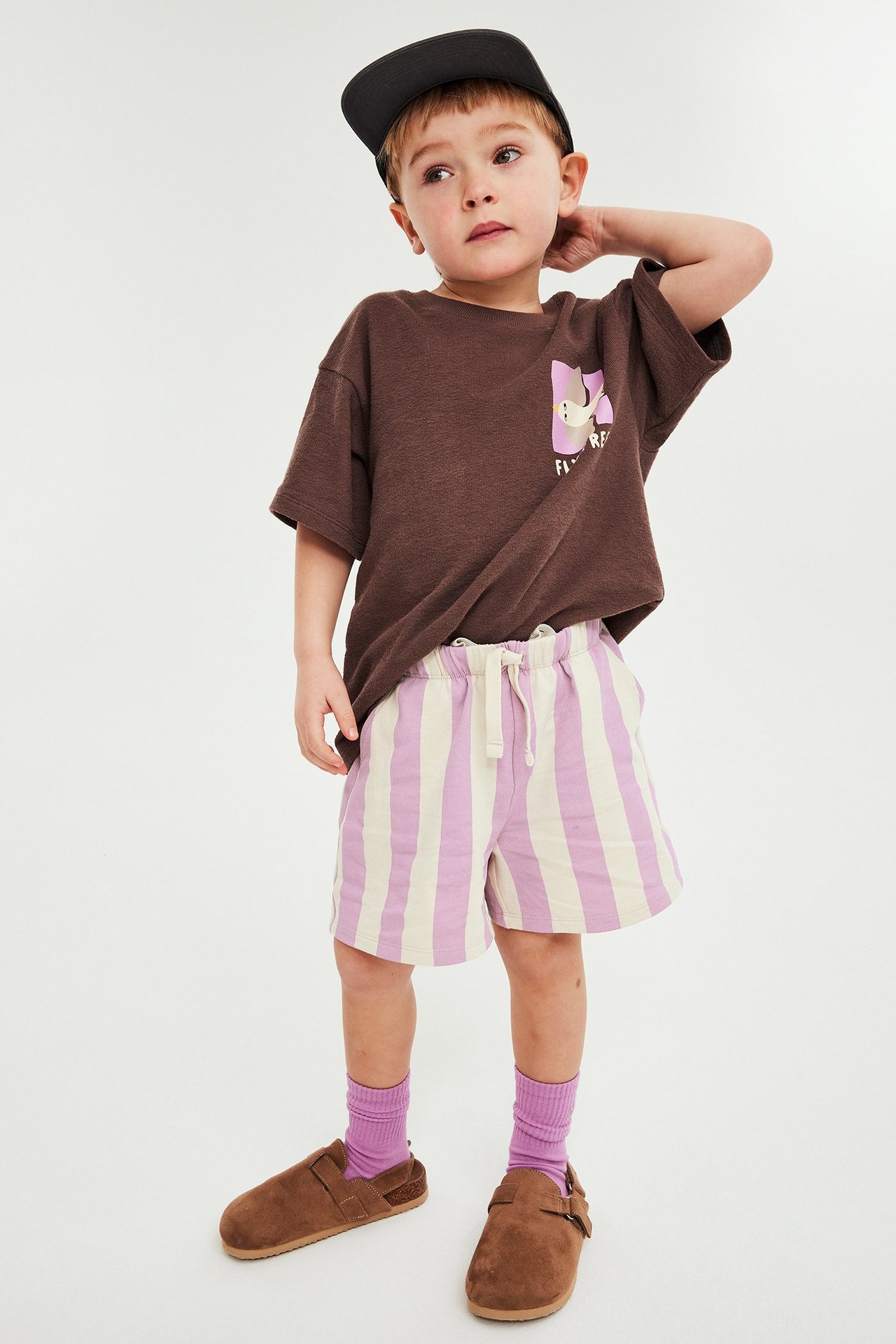 Edestä: Lapsi vaaleanlila/luonnonvalkoisissa raitashortseissa, ruskeassa t-paidassa, mustassa lippiksessä, liloissa sukissa ja tohveleissa.