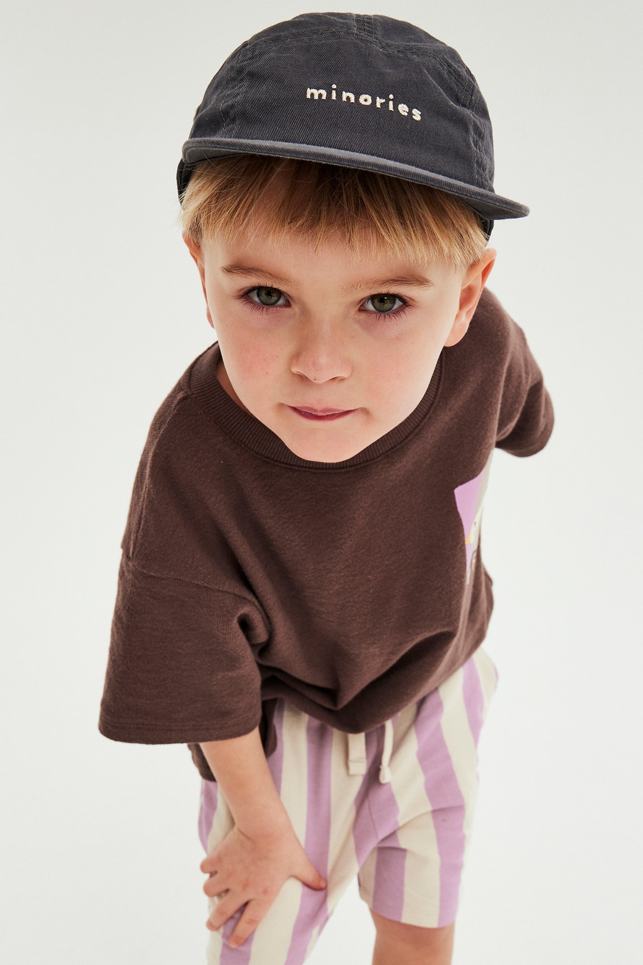 Lapsi yllään vaaleanpunaiset- ja valkoiset raidalliset trikooshortsit, ruskea t-paita ja tummanharmaa lippis.