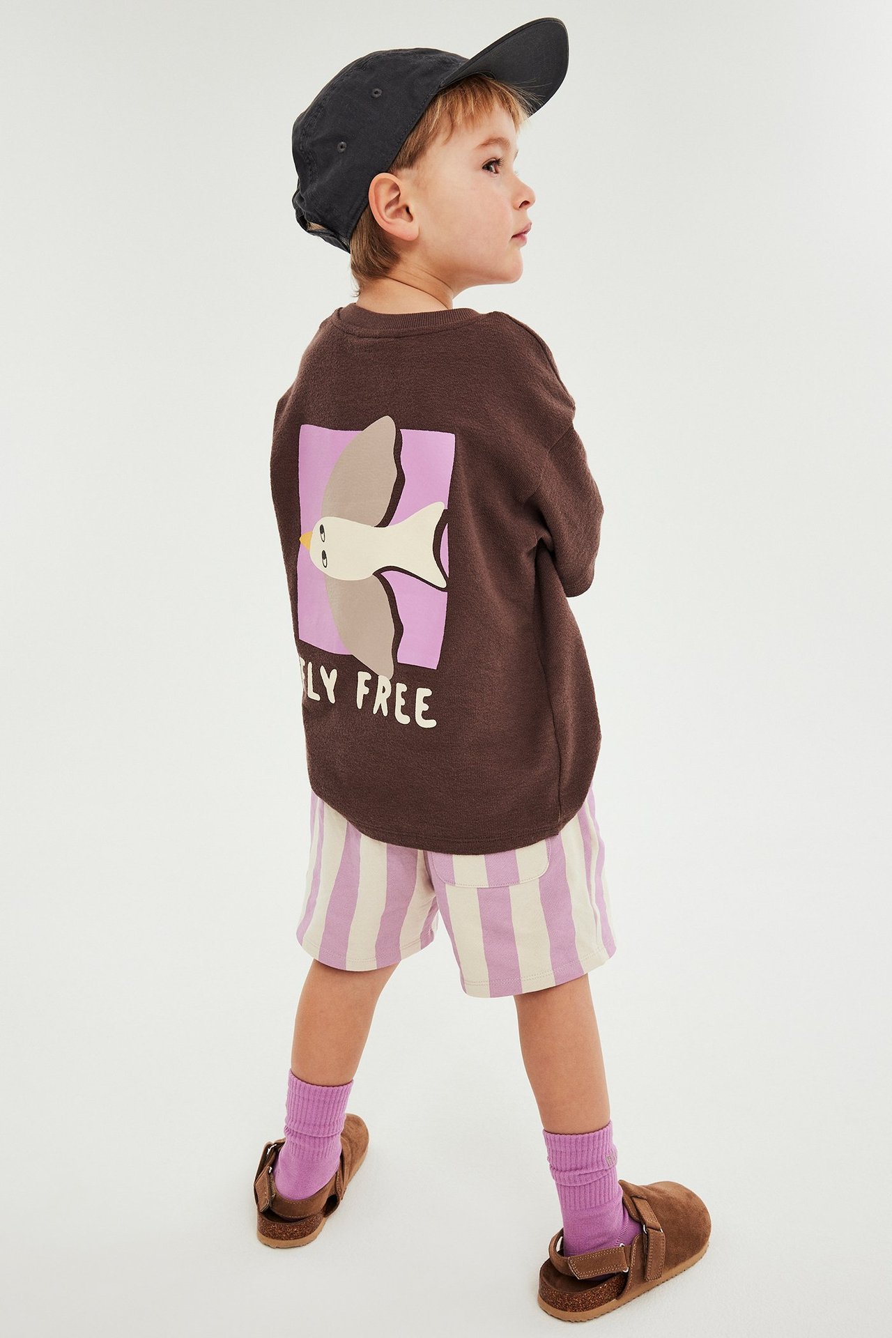 Lapsi, sivukuva: pinkit ja valkoiset raitashortsit, ruskea paita, tumma lippalakki, liilat sukat, sandaalit.