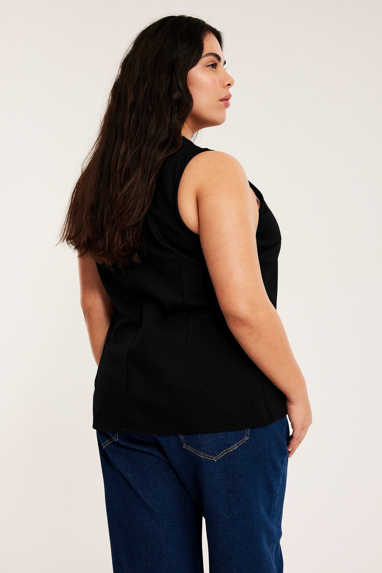 Widok z boku kobiety w czarnej dopasowanej kamizelce z dzianiny i ciemnoniebieskich jeansach.
