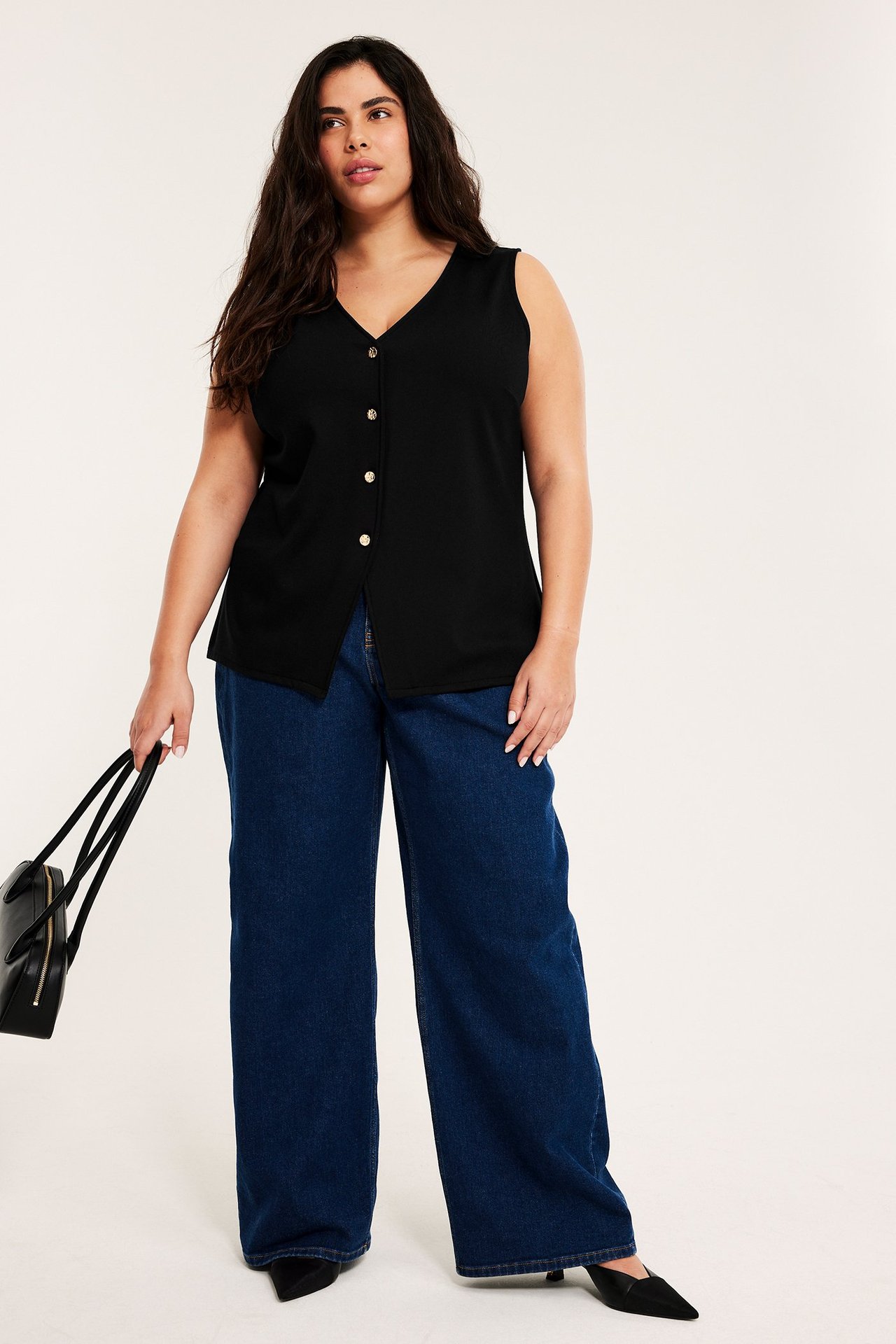 Edestä: Nainen mustassa istuvassa neuleliivissä, tummansiniset farkut, mustat korot ja musta käsilaukku.